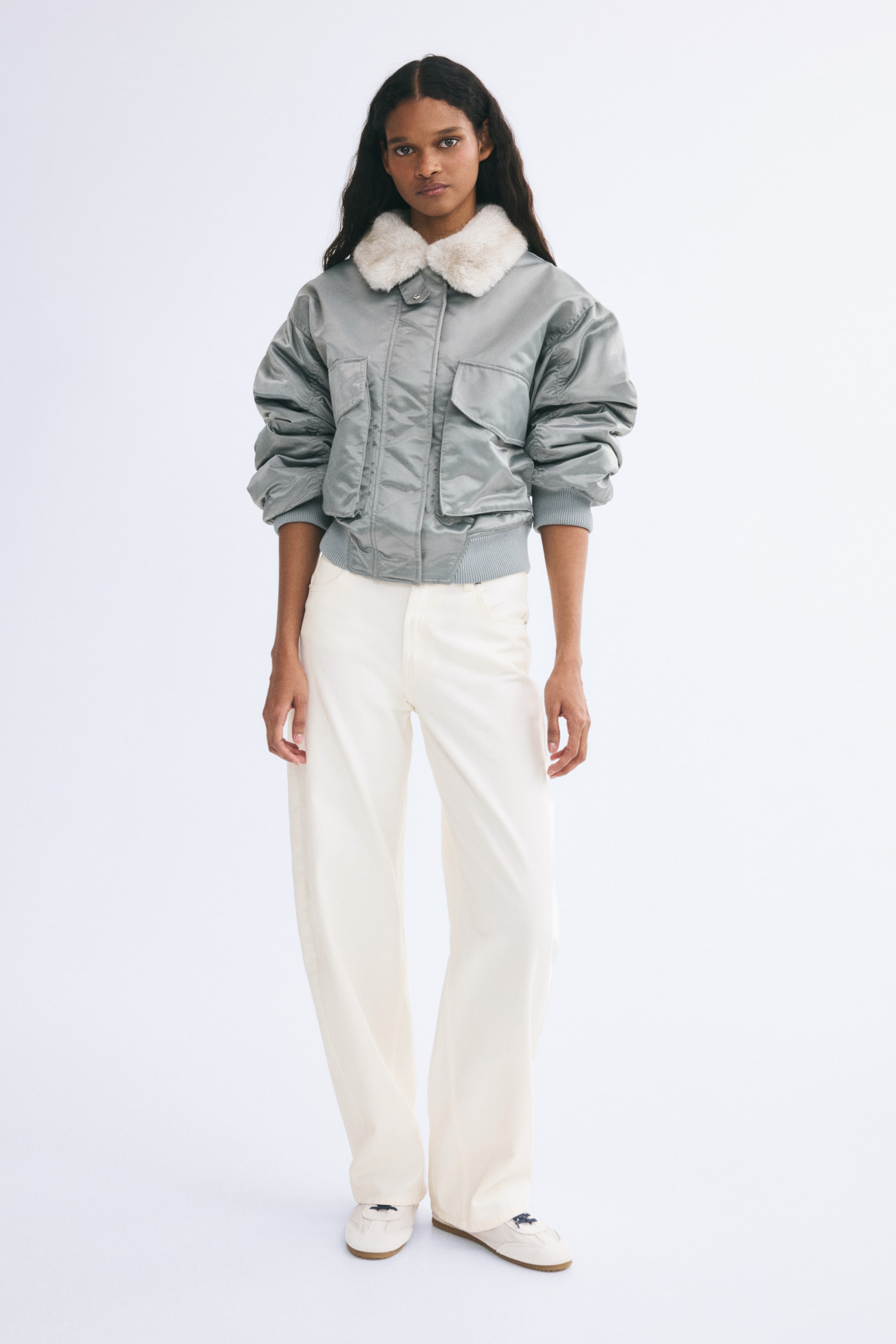 Xem ảnh lớn hơn: Chiếc quần vải twill cotton màu trắng ngà với phom ống rộng cong đang được người mẫu mặc. Quần được phối cùng áo khoác bomber màu xám có cổ lông trắng mềm mại và giày sneaker trắng.