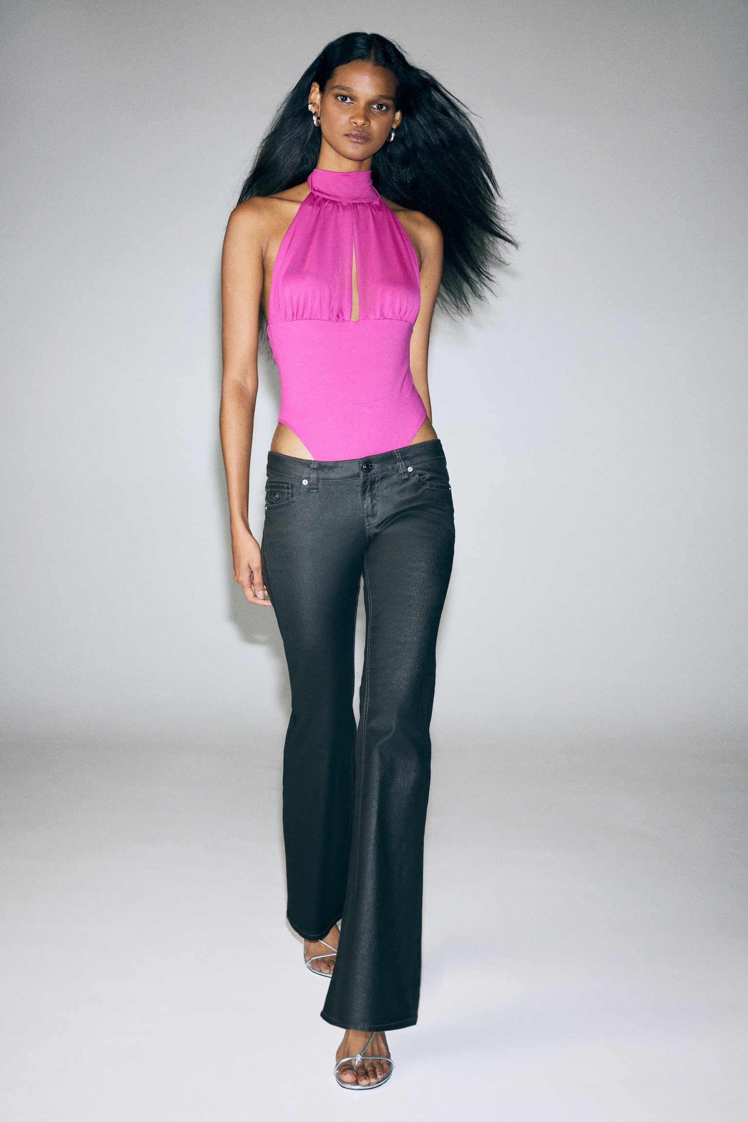 En tettsittende bukse i mÃ¸rk grÃ¥ coated bomullstwill bÃ¦res av modellen, med ekstra lav midje, frontlommer og utsvingte ben. Buksen styles med en livlig rosa halterneck-bodysuit og sÃ¸lvfargede sandaler med remmer. - 1