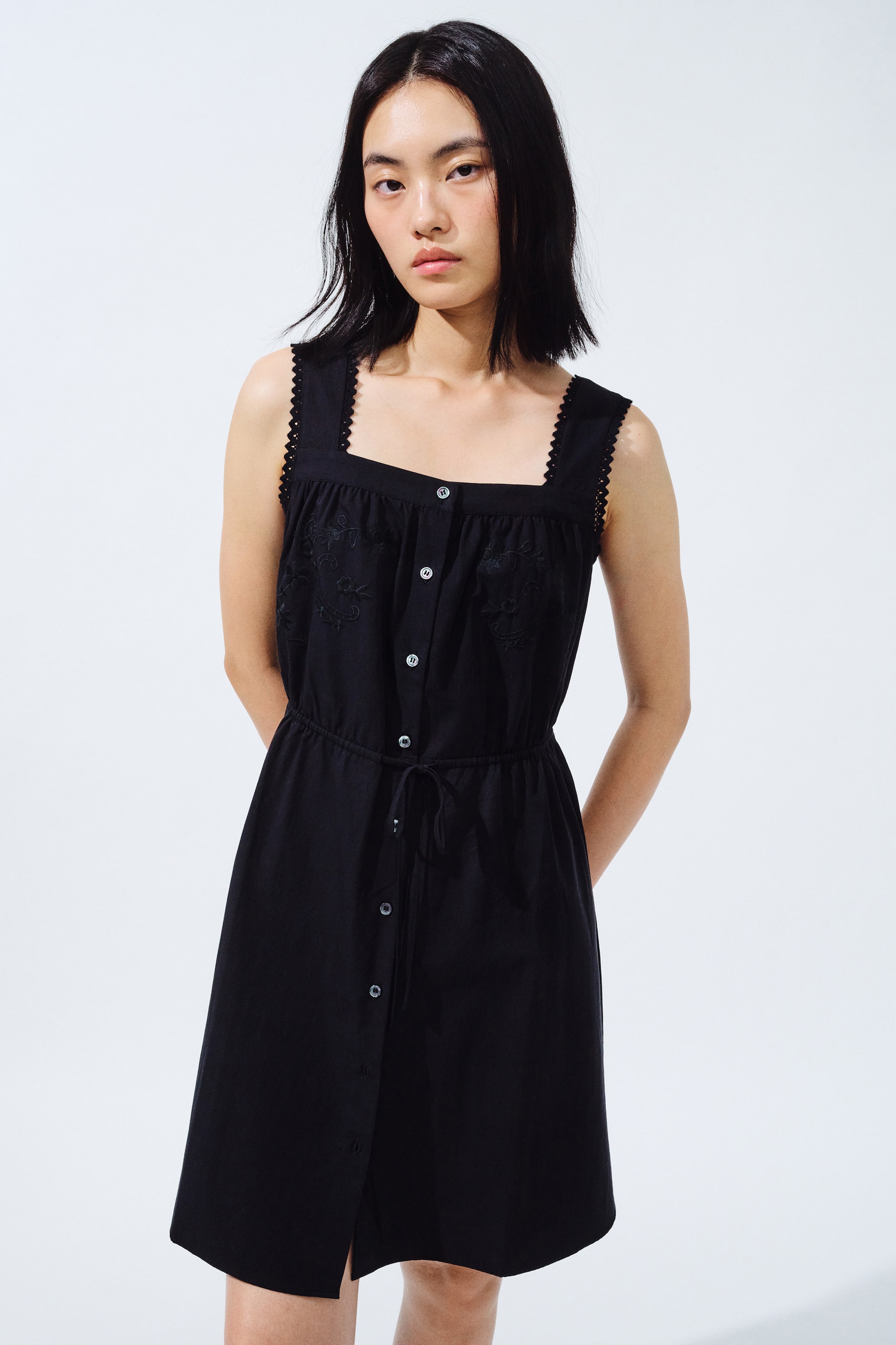View larger image: The black sleeveless dress features a square neckline with scalloped trim on the straps, embroidered detailing on the bodice, and a button-front closure. Designed with a drawstring waist, its skirt falls to approximately knee length, offering a front-facing view.