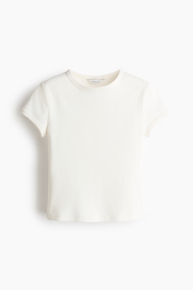 Ein cremeweißes T-Shirt präsentiert einen Rundhalsausschnitt und kurze Ärmel mit umgeschlagenen Bündchen, von vorne gesehen. Es zeichnet sich durch eine glatte Stoffstruktur, einen geraden Saum und ein kleines Markenetikett aus, das am hinteren Hals sichtbar ist. - 1