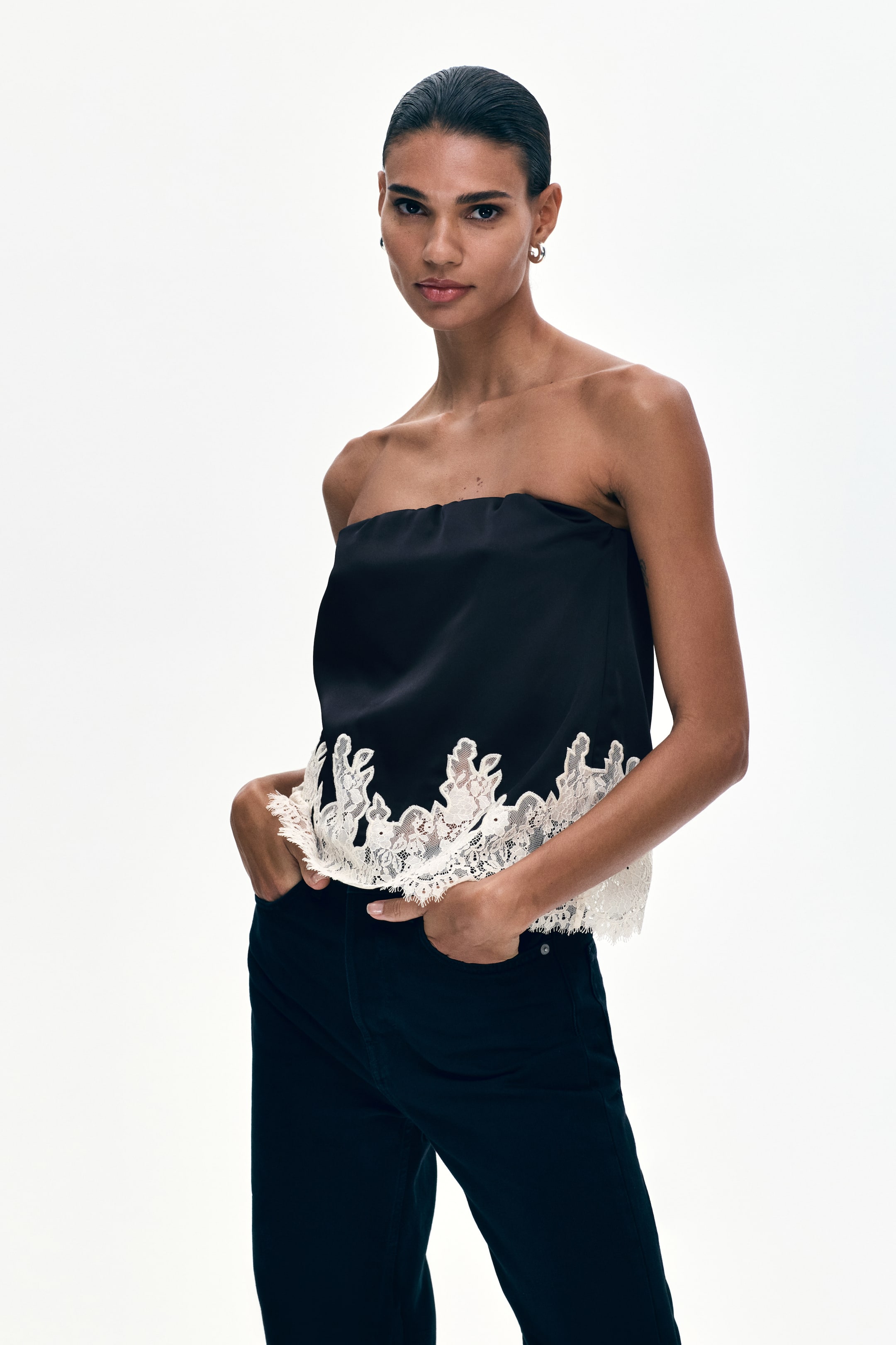 View larger image: The model is wearing a black strapless top. An off-white lace trim with a scalloped edge adorns the bottom hem of the top. The model is placing her hands in the pockets of dark trousers.