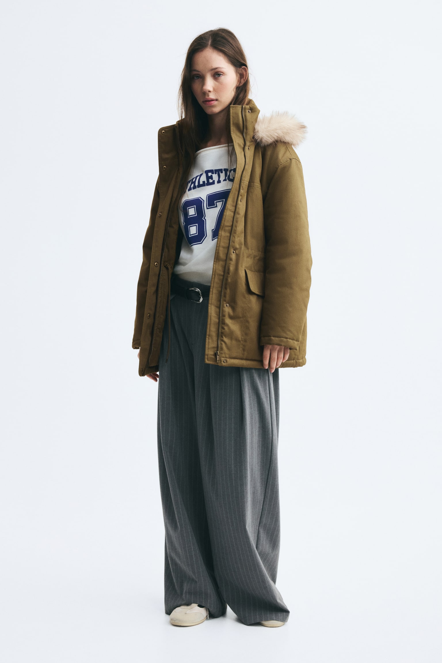 An oversized, lightly padded olive green parka is worn open by the model, featuring a hood with fluffy faux-fur trim and front flap pockets. The parka is layered over a white graphic sweatshirt and wide-leg, pinstriped grey trousers. - 1