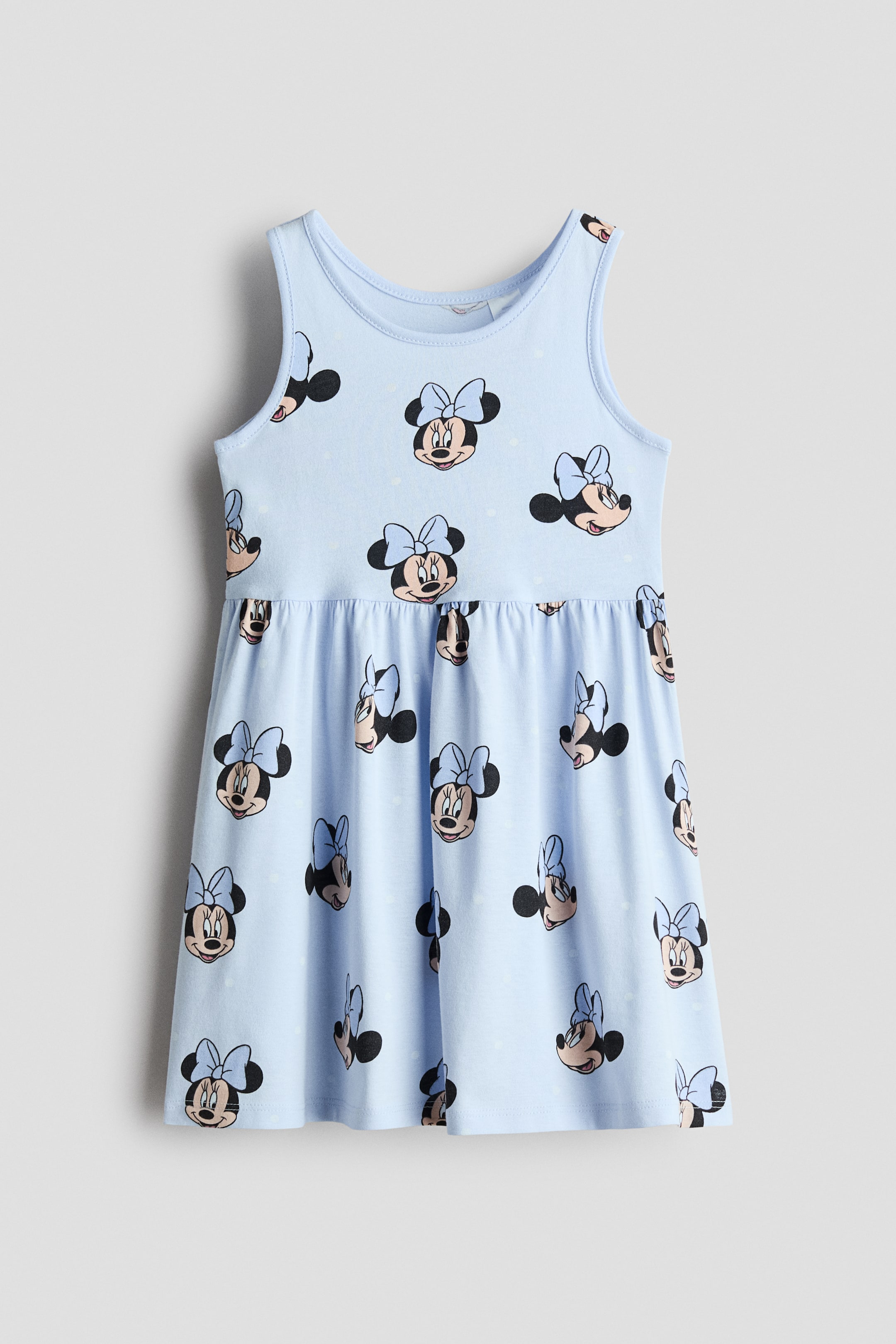 View larger image: A light blue sleeveless dress is shown front-facing, featuring an all-over print of Minnie Mouse faces with light blue bows. The dress has a round neckline, a gathered seam at the waist, and a flared skirt.