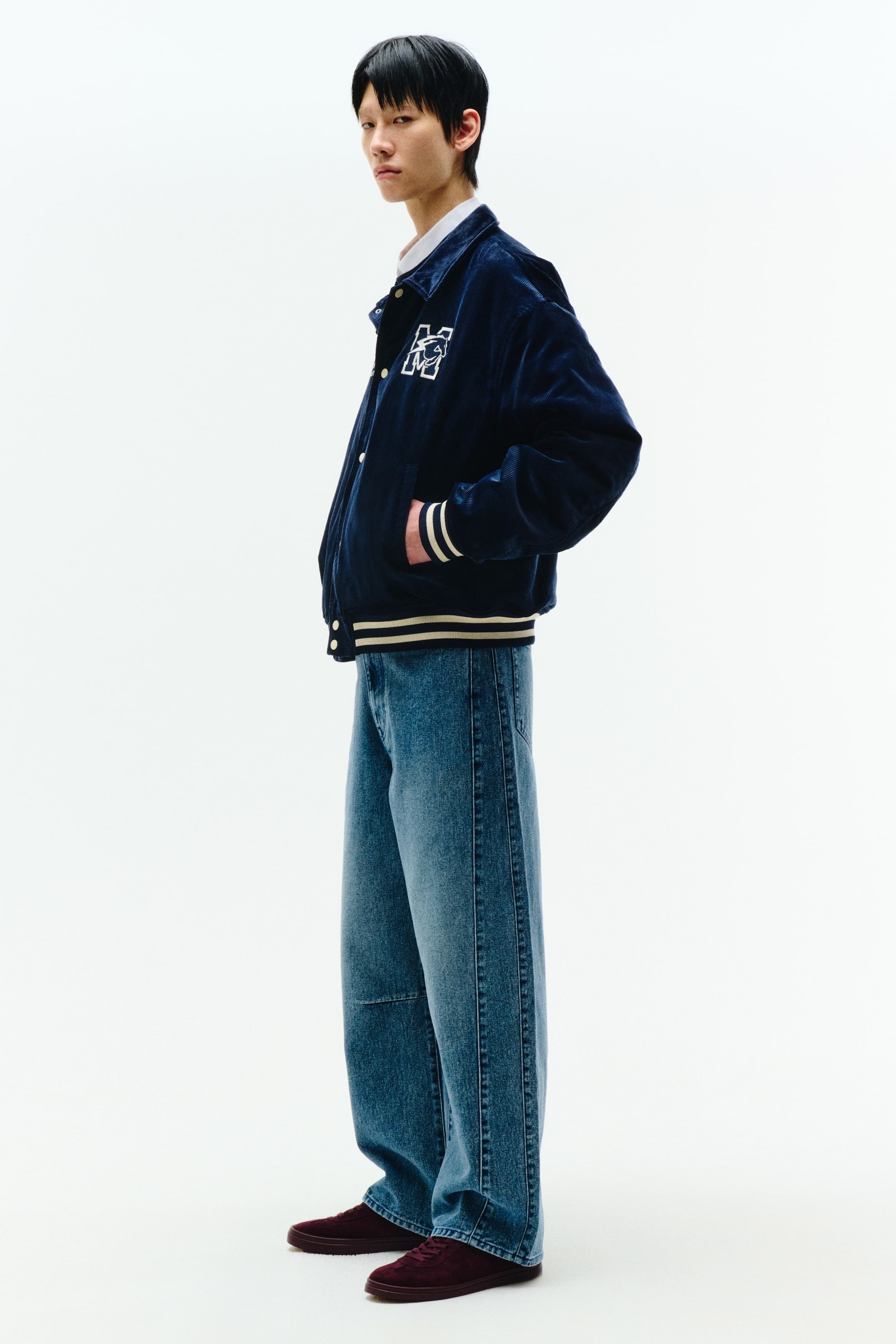 拡大画像を表示: ネイビーのコーデュロイバルマカンジャケットは、白い襟付きシャツの上に着用され、そのサイドプロファイルを披露しています。それは、尖った襟、大きな白の「M」ロゴが左胸に卒業帽と共に配置され、スナップボタン式のクローザー、そしてストライプのリブ付きカフスと裾が特徴です。