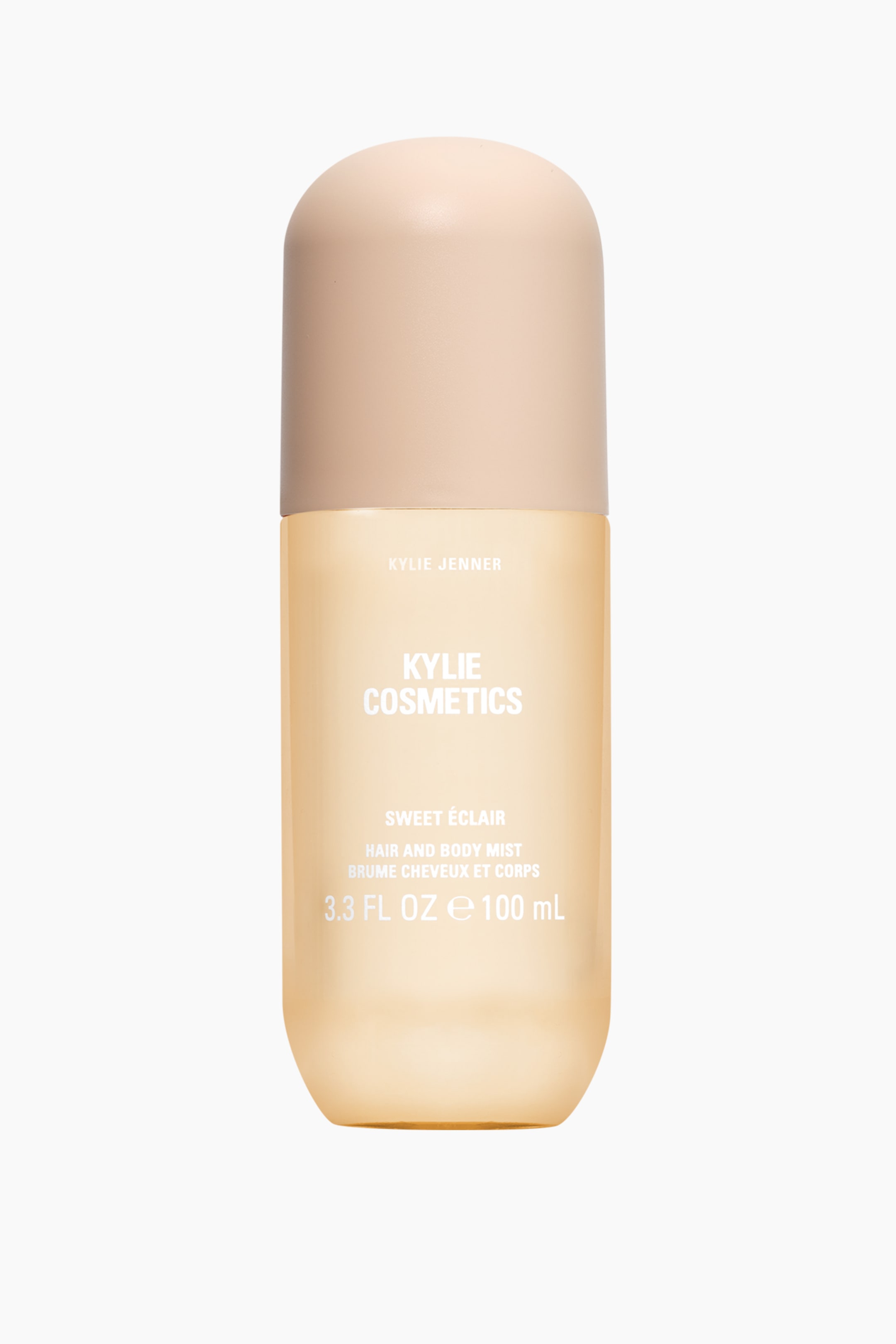 Se større billede: En Kylie Cosmetics Sweet Éclair Hair and Body Mist-flaske står oprejst og vender fremad. Den har en gennemsigtig ravfarvet krop, der diskret smalner mod bunden, og er toppet med en afrundet, mat beige låg. Hvid tekst er trykt på forsiden, herunder "Kylie Cosmetics" og "Sweet Éclair".