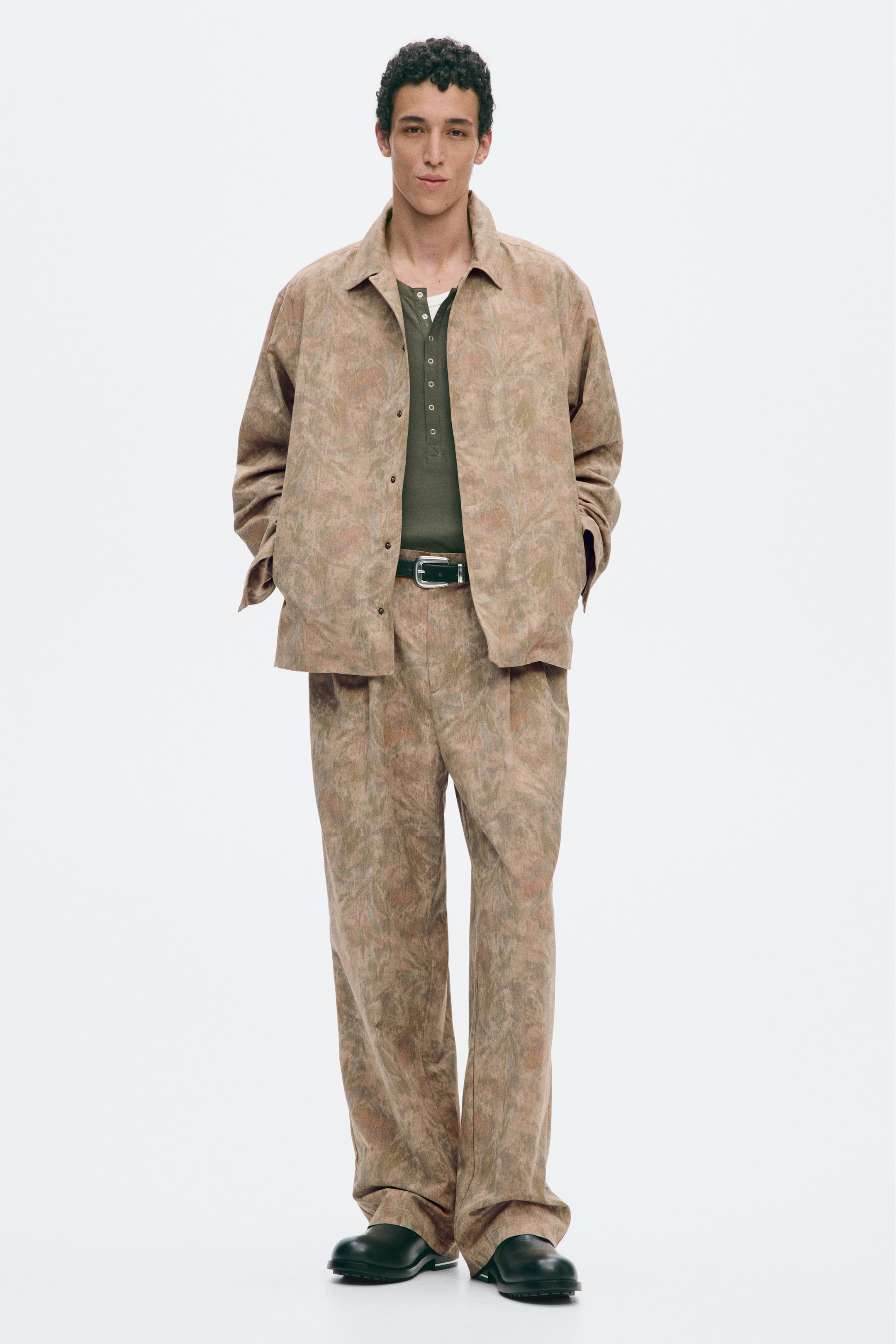 Visualizza immagine più grande: Pantaloni in tessuto intrecciato a fantasia, caratterizzati da una vestibilità rilassata, gamba dritta e pieghe frontali, indossati dalla modella. I pantaloni sono abbinati a una giacca aperta coordinata, a una camicia verde scuro infilata nei pantaloni e fermata da una cintura nera, e a scarpe nere.