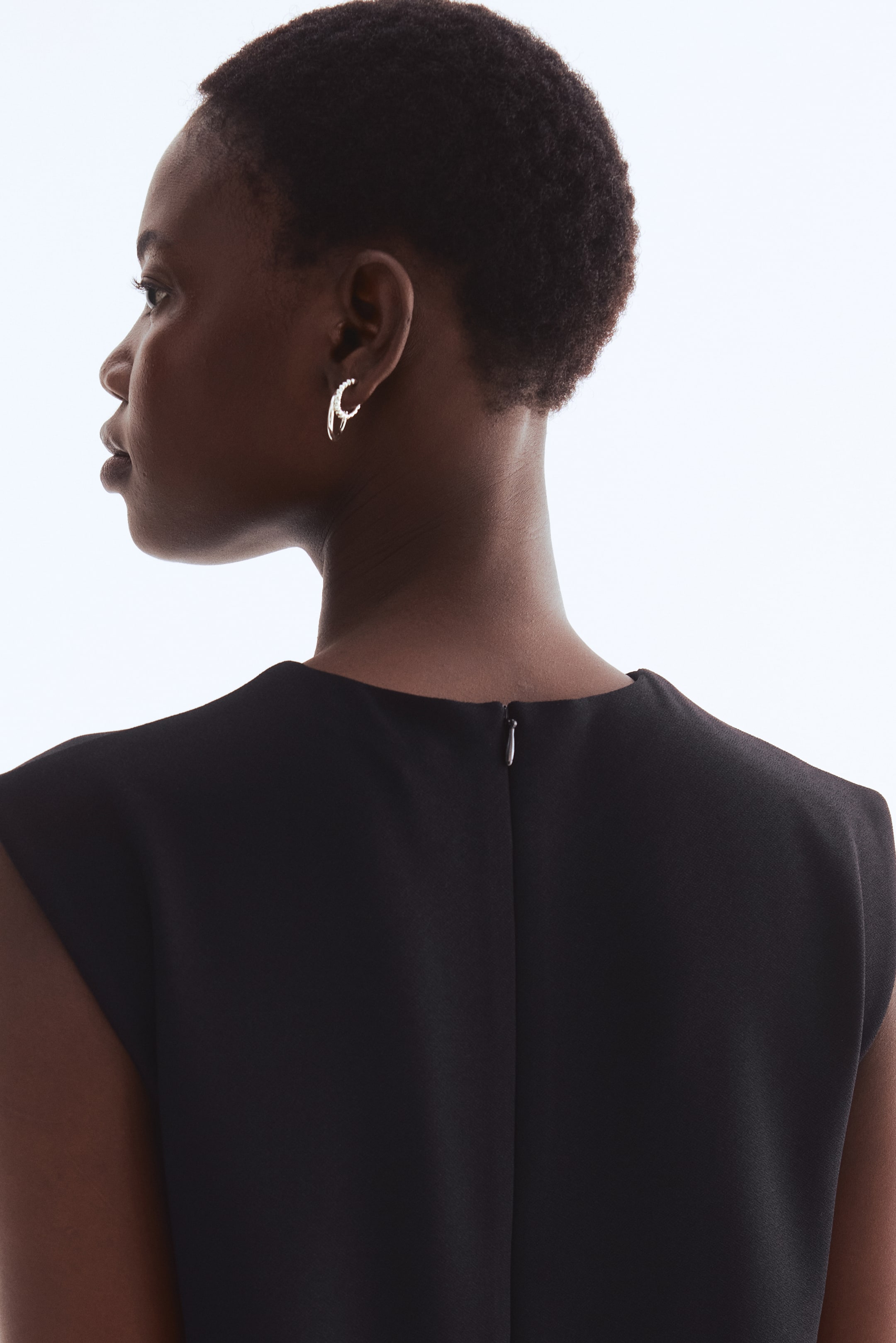 Se større billede: Sølvtone dobbelte bøjleøreringe, hvor den ene bøjle er enkel og den anden pyntet med små sten, bæres fra en sideprofil. Et sort, ærmeløst klædningsstykke præsenteres fra ryggen, der viser en høj halsudskæring og en synlig sølv lynlåslukning.