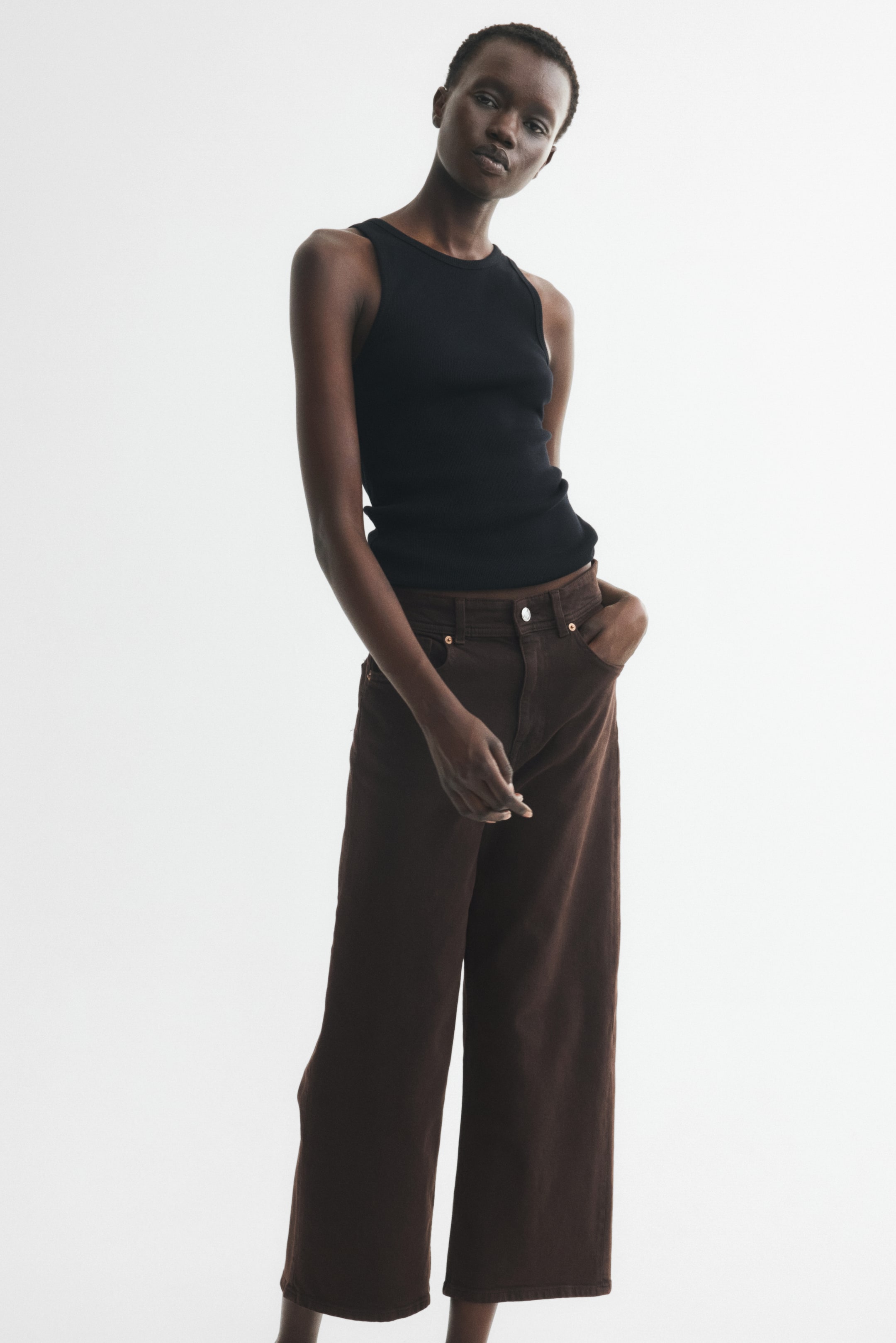View larger image: Dark brown 5-pocket jeans with a high waist and button-and-zip fly closure are being worn by the model, who is resting one hand in a front pocket. The jeans feature a wide cut from the thigh to the ankle.