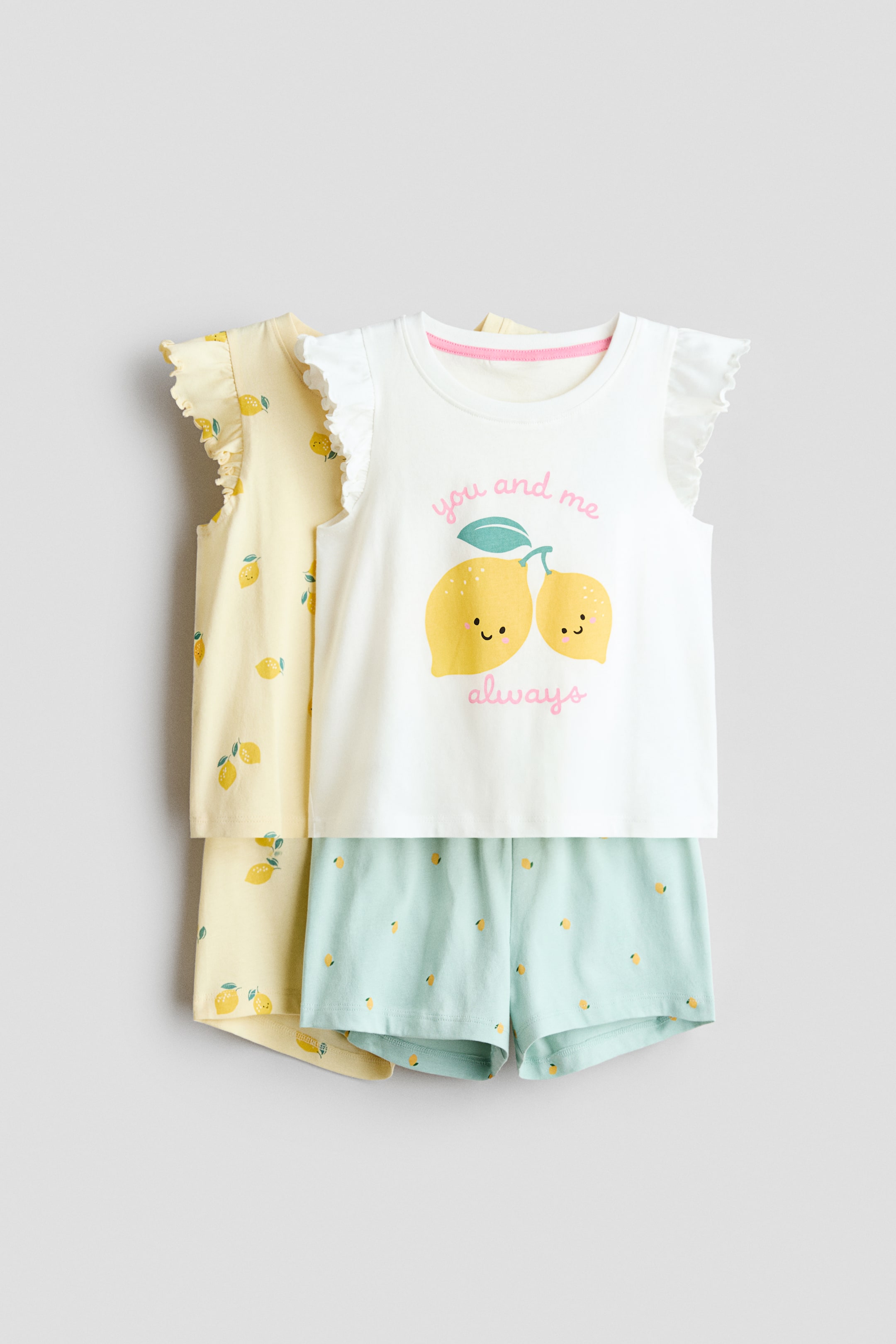 查看大圖: 柔软棉质针织睡衣以两套形式呈现,前套清晰可见为白色上衣和浅蓝色短裤。白色上衣具有圆形罗纹饰边领口、飘逸袖,并在中央印有两个微笑柠檬的图案,而浅蓝色短裤则展示了松紧腰带和小柠檬印花;后面部分可见的黄色套装则是满印柠檬图案。