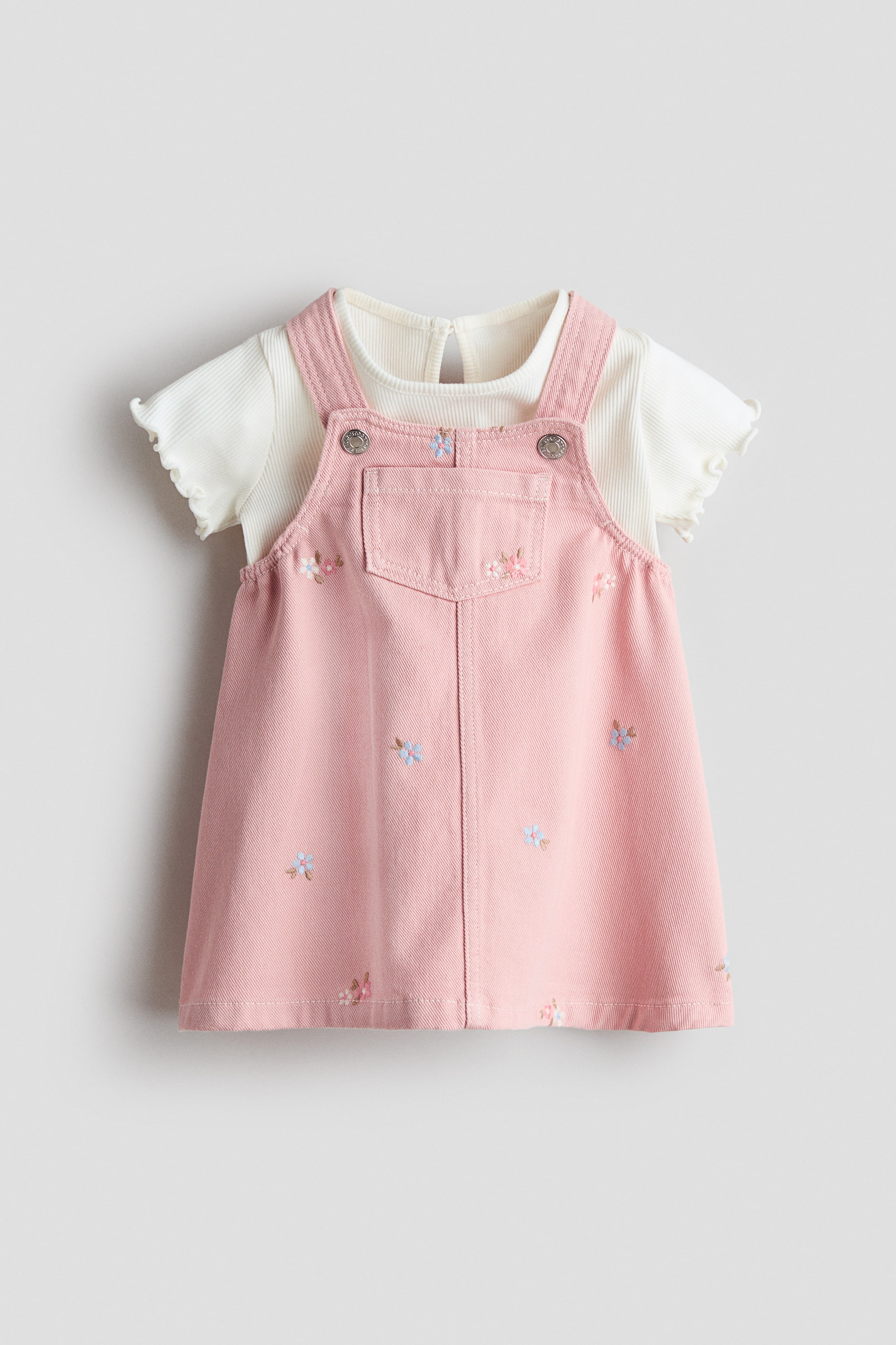 Ver imagen más grande: Conjunto de dos piezas para bebé visto desde el frente, compuesto por un vestido de peto rosa sobre una camiseta de canalé color crema. El vestido de peto rosa presenta un escote cuadrado, tirantes ajustables con corchetes, un bolsillo abierto en el pecho y bordados florales dispersos, mientras que la camiseta de manga corta color crema muestra un escote redondo y puños con volantes.