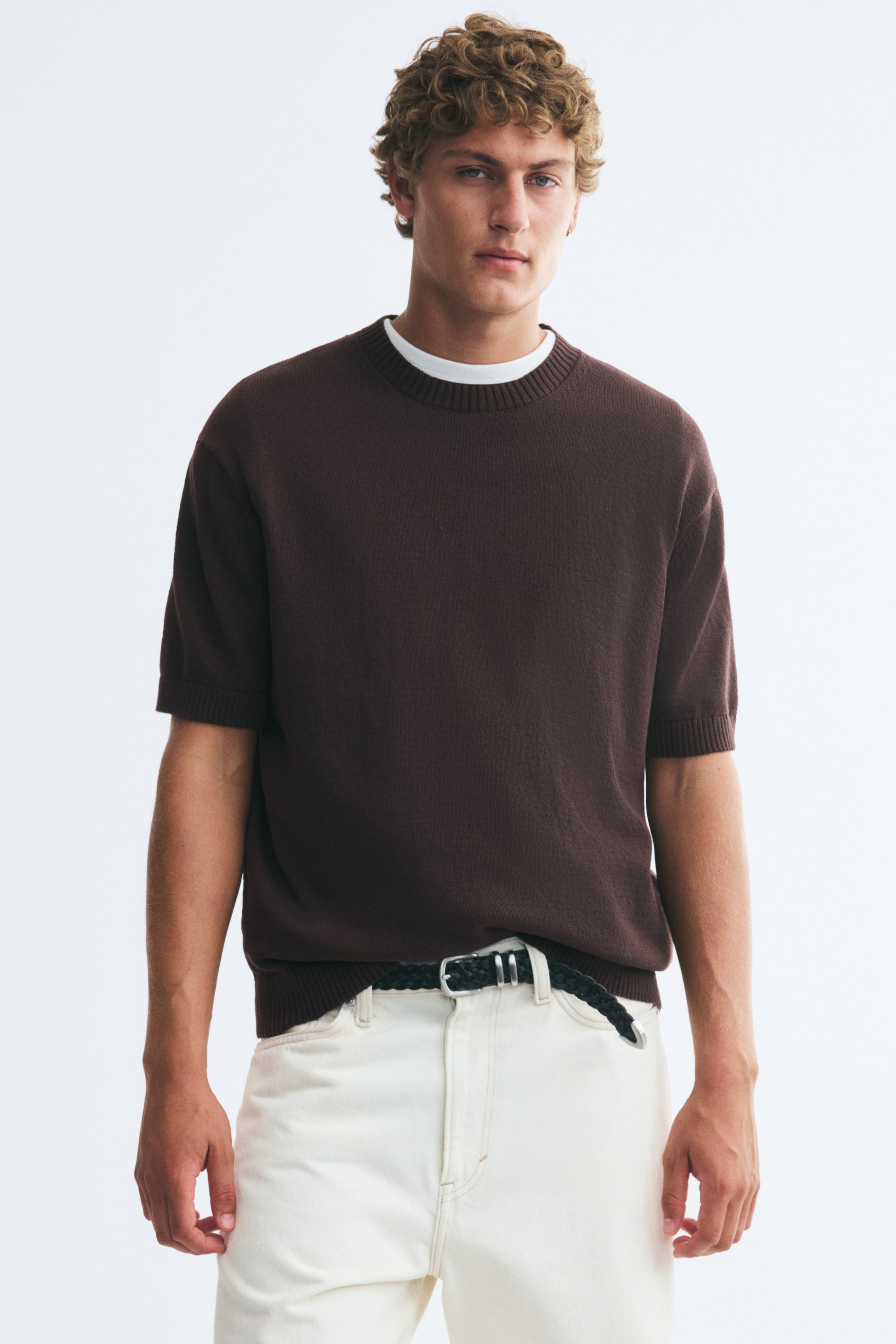 View larger image: The model is wearing a dark brown knitted T-shirt, featuring a regular fit, a ribbed crew neckline, and ribbed cuffs and hem. The T-shirt is layered over a white undershirt and paired with light-colored trousers.