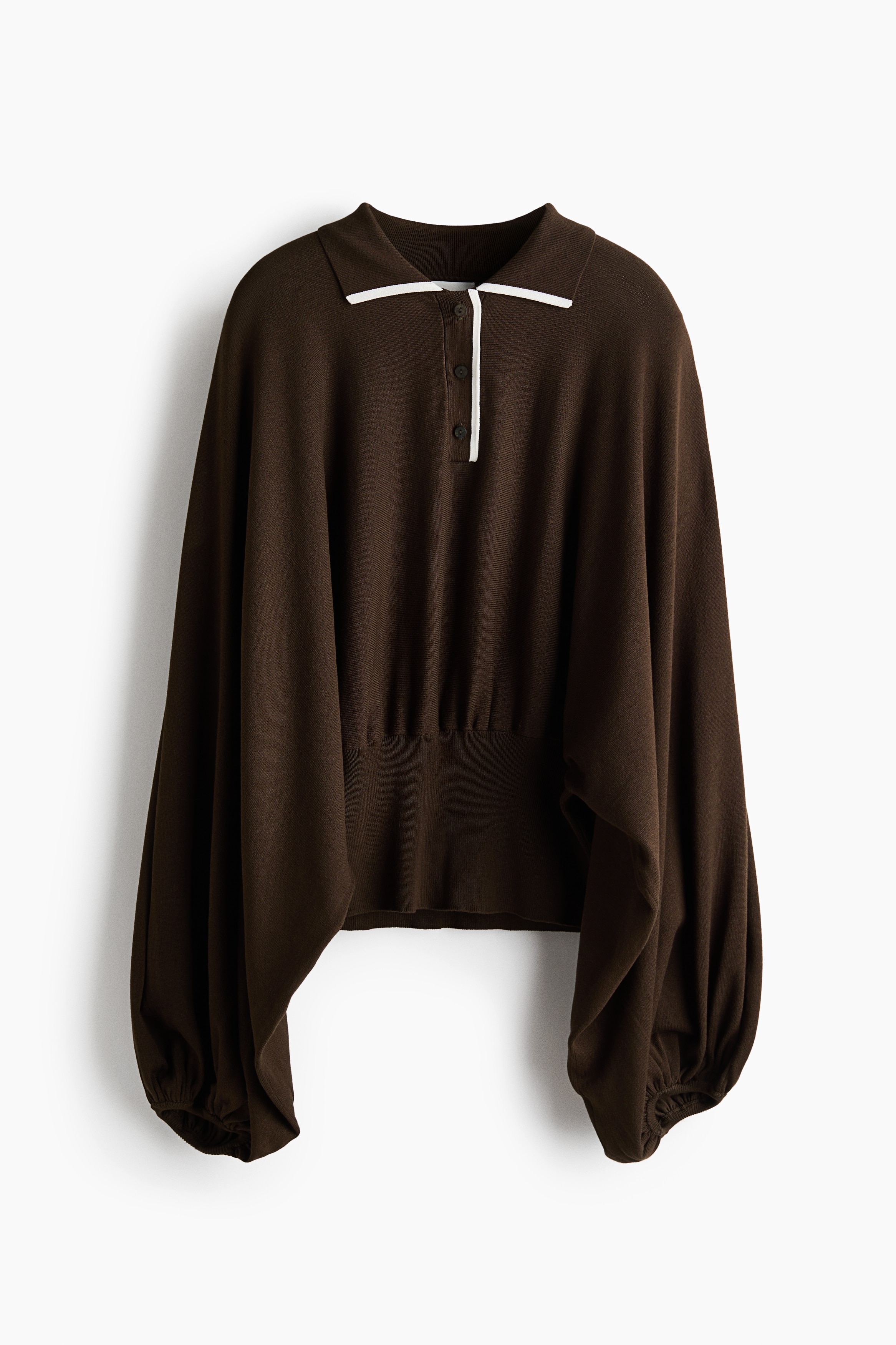 The model is wearing a brown knit polo sweater, featuring a white-trimmed collar, button placket, and long, voluminous batwing sleeves. The sweater has a wide, fitted waistband with gathers along the top edge and is paired with white jeans.