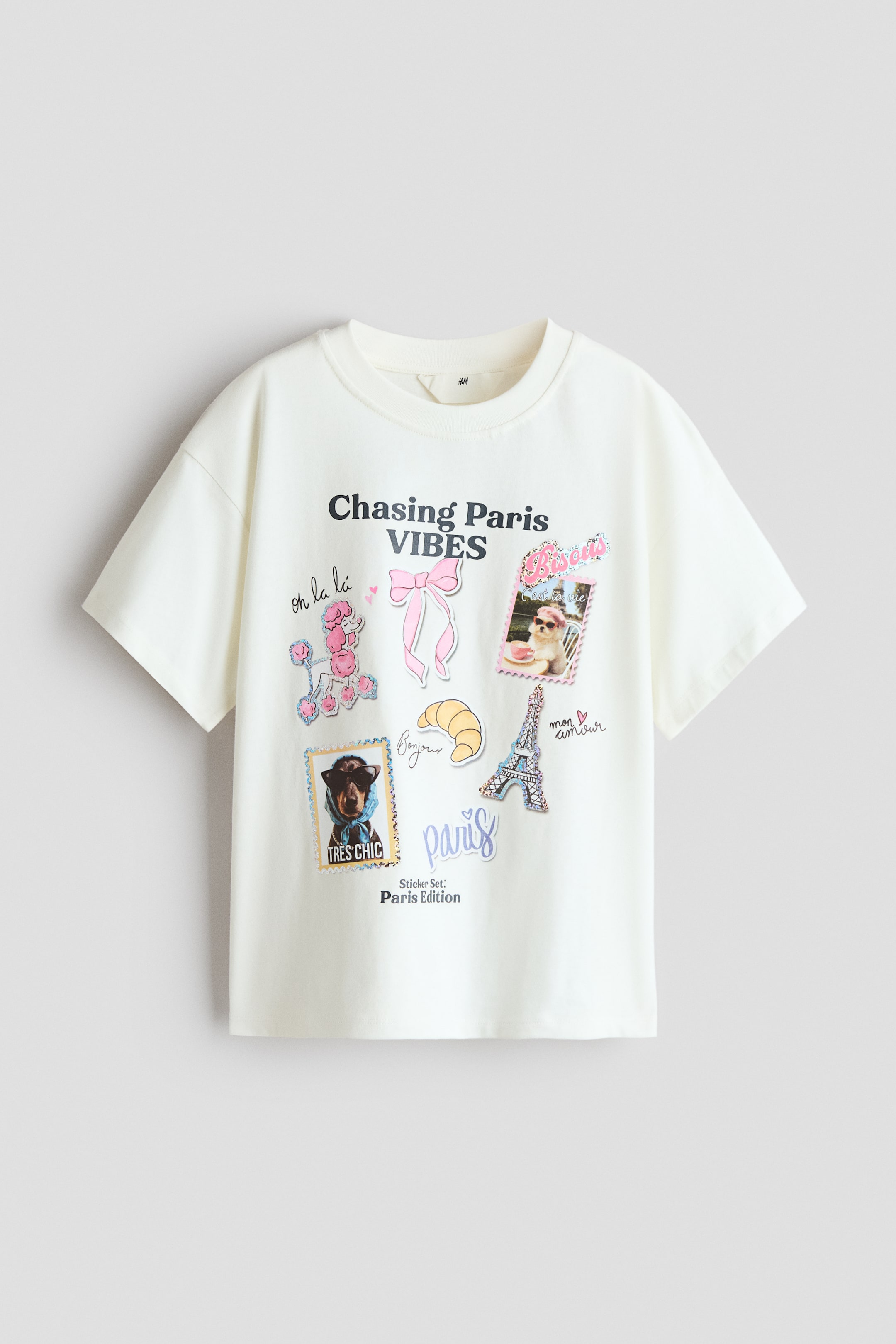 Prikaži veću sliku: Široka, prljavo bela majica od mekog, štampanog pamučnog dresa okrenuta je napred, prikazujući okrugli izrez sa rebrastim obrubom i spuštena ramena. Na prednjoj strani ima šareni grafički print sa raznim ilustracijama i tekstovima na temu Pariza, uključujući 'Chasing Paris VIBES', ružičastu mašnu i Ajfelovu kulu.