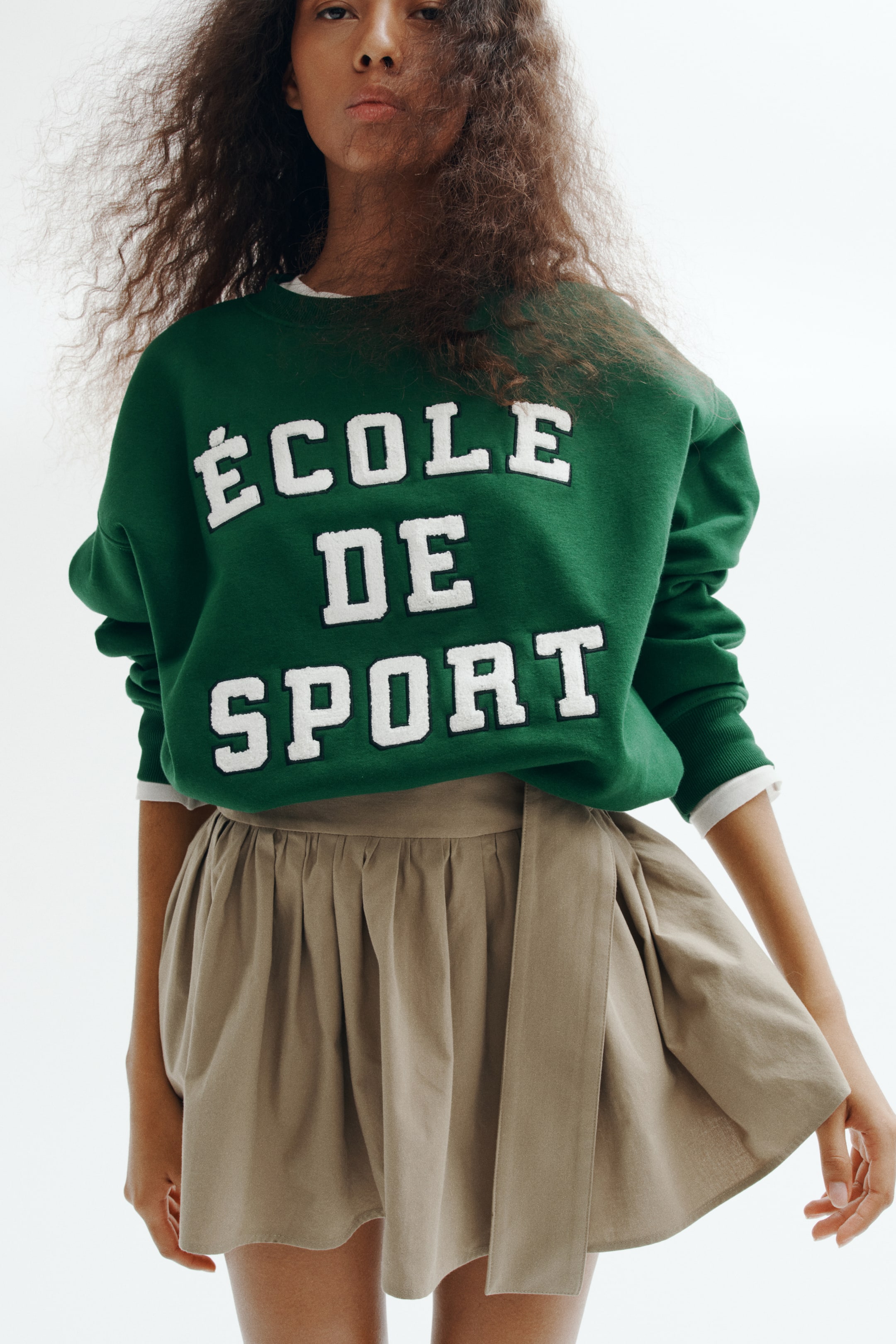 View larger image: An oversized green sweatshirt featuring a white text design that reads "�COLE DE SPORT" is worn by the model. The sweatshirt, designed with a ribbed crew neckline and dropped shoulders, is layered over a white top and styled with a beige flared skirt.