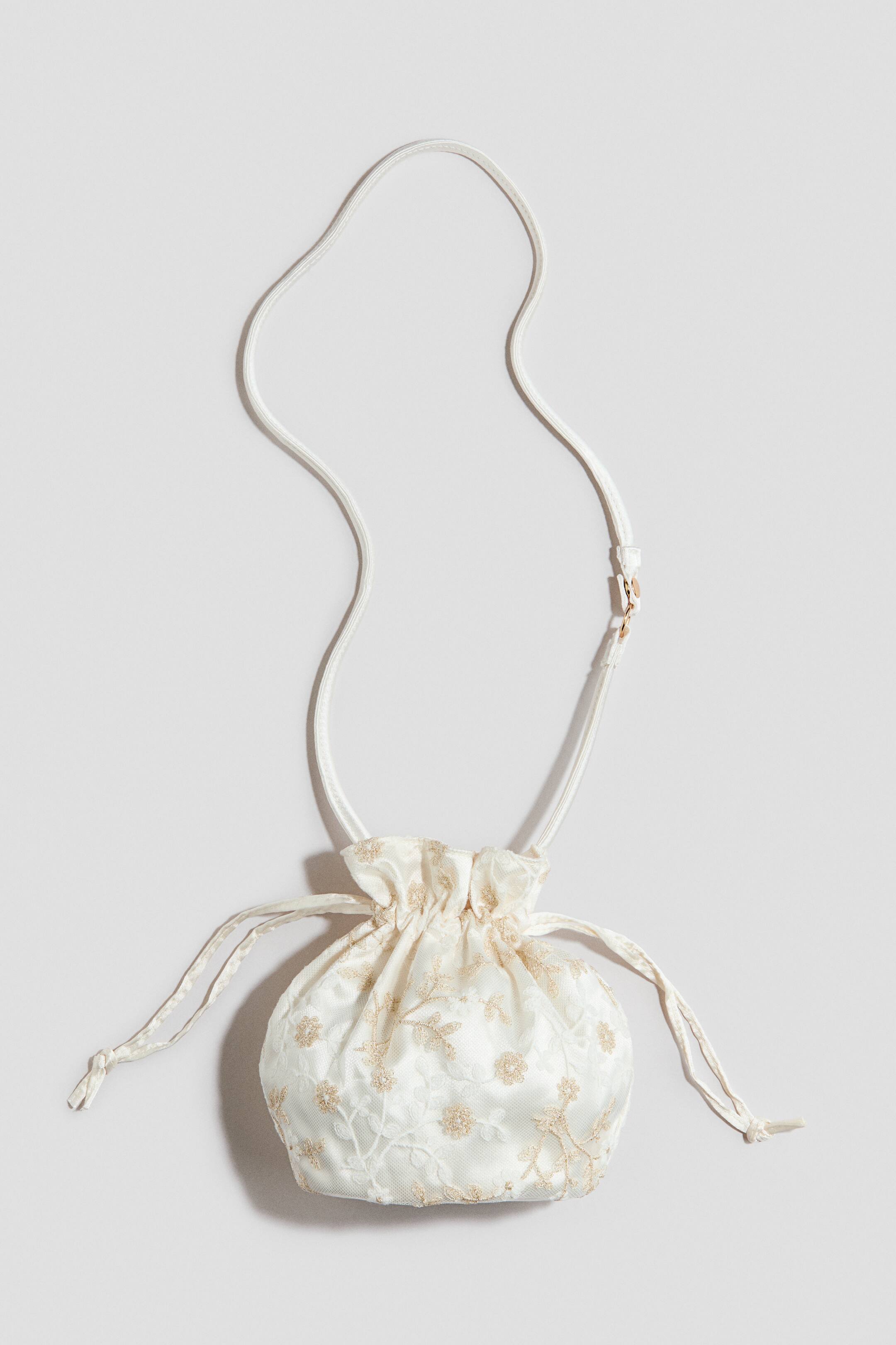 הצגת תמונה גדולה יותר: תיק כיס לבן-מחוץ-לבן עם שרוך מוצג בחזית, מעוטר ברקמת פרחים זהובה מעל תחרה שקופה. שרוך תואם מכווץ את החלק העליון, ורצועה דקה ומתכווננת נמתחת מצִידָו.