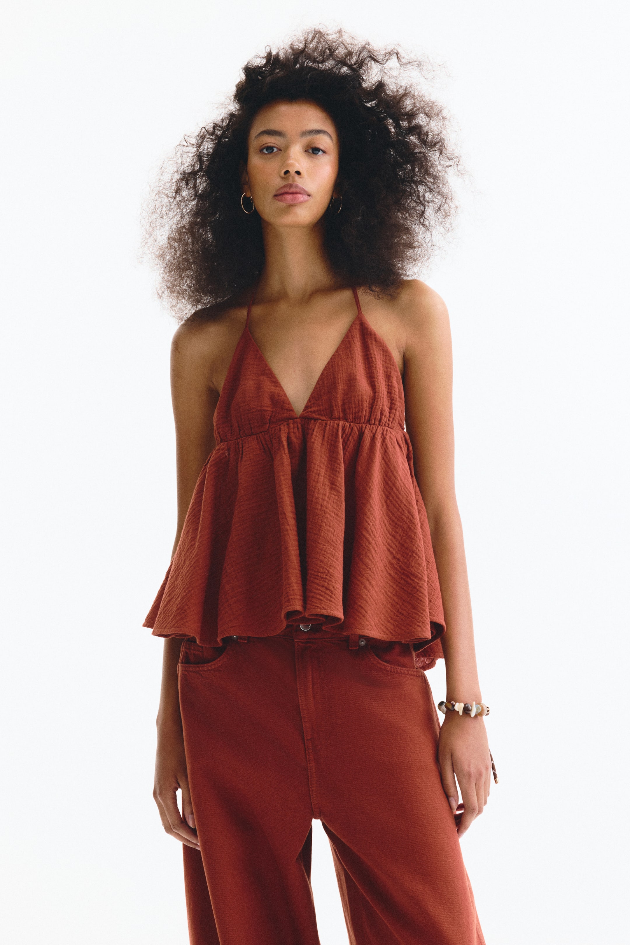 View larger image: A rust-coloured halter neck top showcases a crinkled texture, a deep V-neckline, and a gathered peplum hem, creating a flowing silhouette. The top is facing forward and is styled with matching wide-leg trousers.