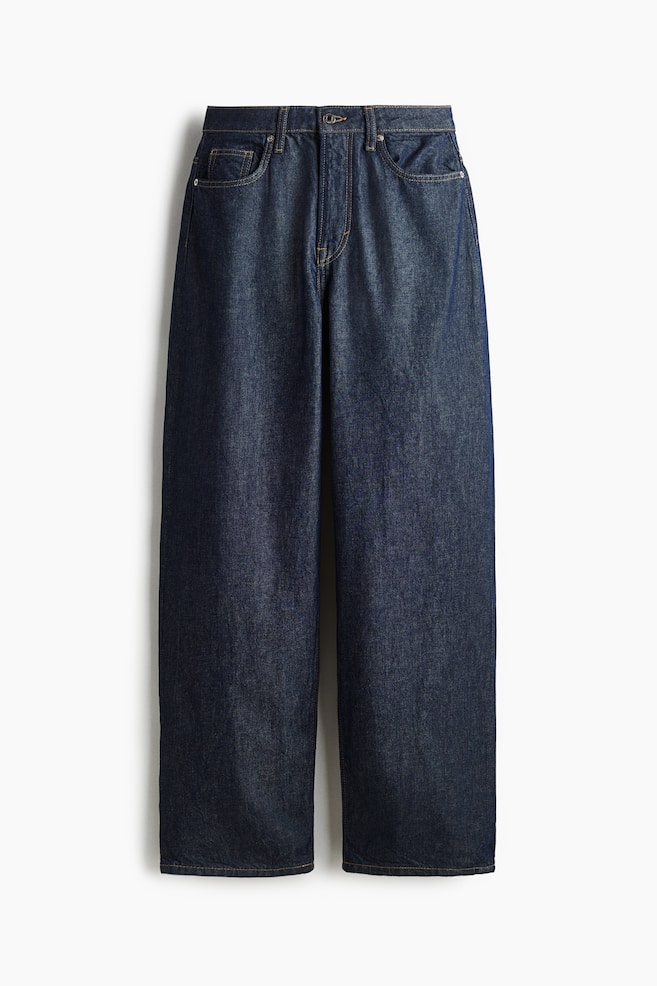 Un paio di jeans in denim di cotone blu scuro a 5 tasche è esposto FRONTE, caratterizzato da una vita alta con chiusura a zip e bottone. I jeans sono progettati con una gamba curva e voluminosa, offrendo una vestibilità ampia dall'anca alla coscia e una vestibilità larga e affusolata dalla coscia all'orlo. - 1