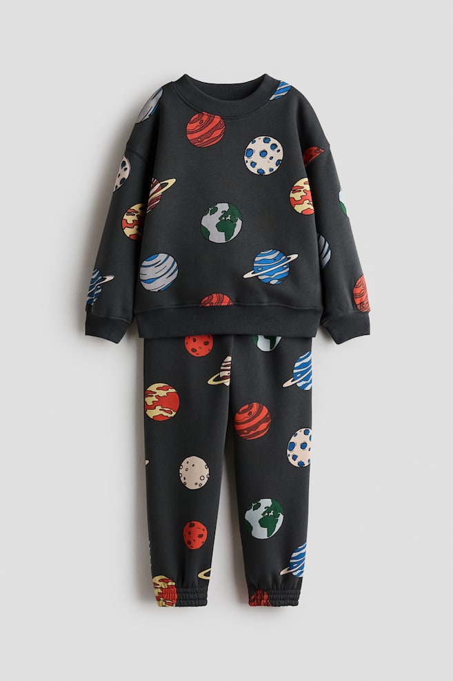 A matching dark grey sweater and track pants set is displaying from the front, featuring an all-over colorful planet print. The long-sleeve sweater is showcasing dropped shoulders, a ribbed crew neck, and ribbed cuffs and hem, while the matching track pants are featuring elasticated ankle cuffs. - 1