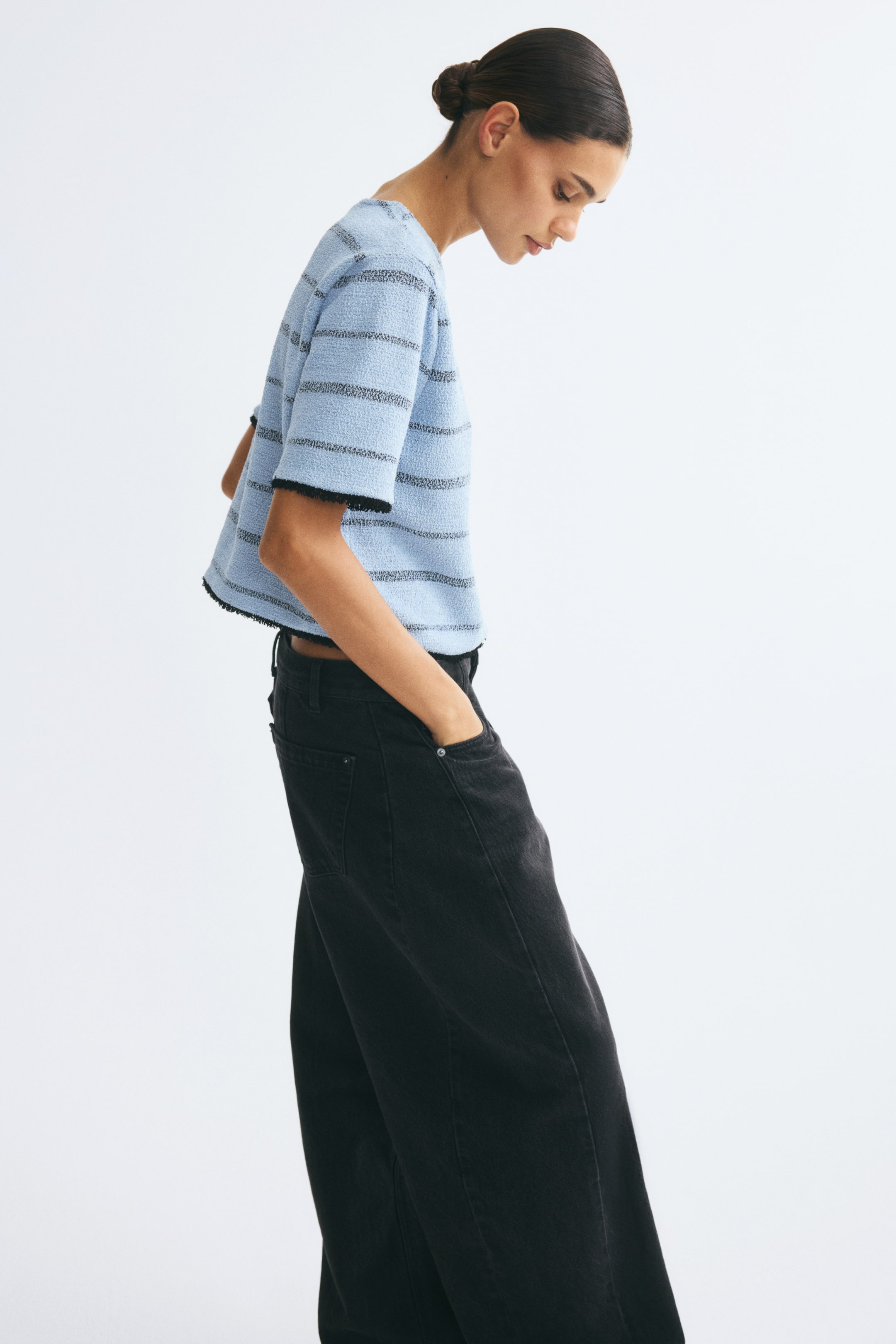 Grotere afbeelding bekijken: Het model draagt een kort, boxy topje van lichtblauwe gestructureerde jersey met horizontale zwarte strepen, terwijl ze met haar handen in de zakken van een wide-leg zwarte jeans staat. Het topje heeft een ronde hals, korte mouwen en zwarte franjebies langs de manchetten en de zoom.