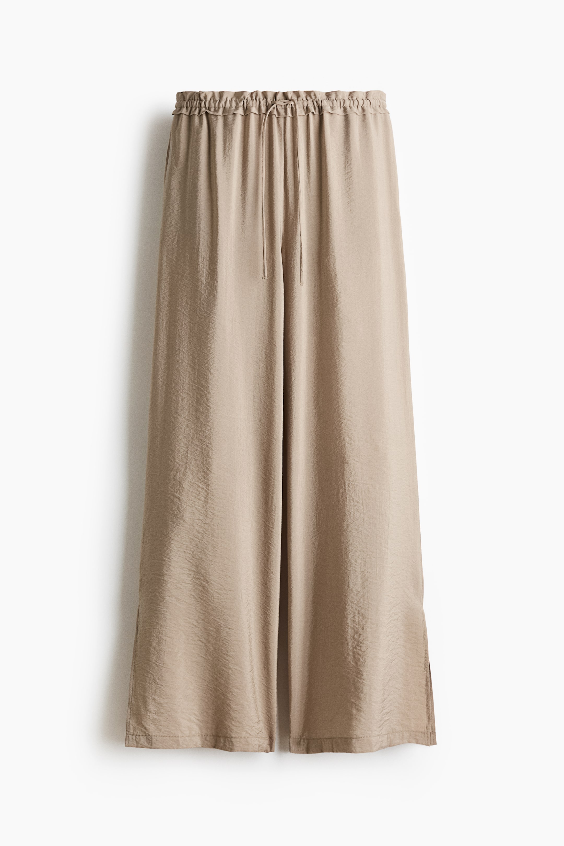 Grotere afbeelding bekijken: Beige wijde broek met een elastische tailleband met zichtbare trekkoord aan de voorkant. De broek heeft een gekreukelde textuur en kleine zijsplitjes aan de zoom.