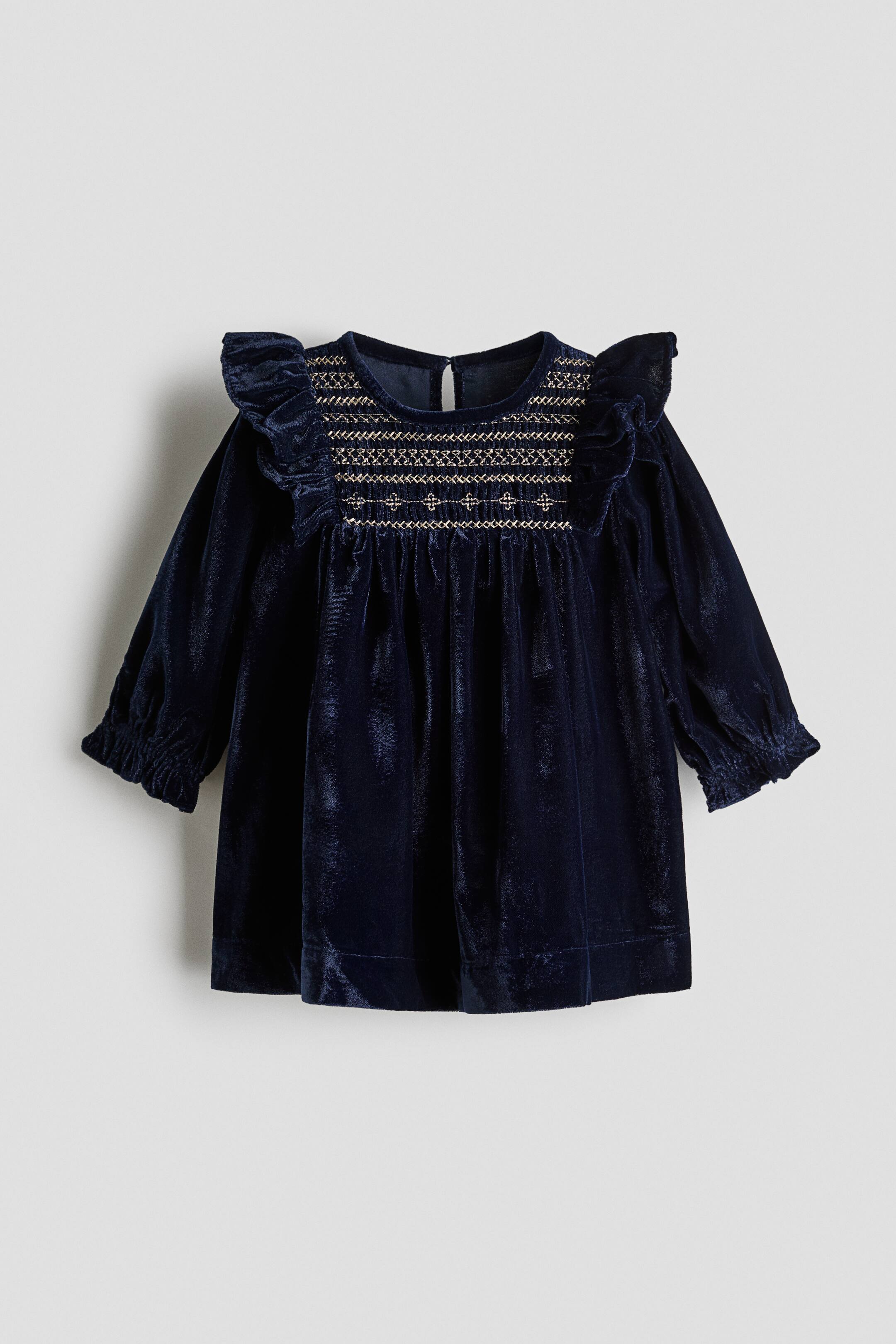 Agrandir l'image: Une robe bébé en velours bleu marine est présentée de face, mettant en valeur un plastron smocké orné de motifs brodés dorés et de finitions à volants aux épaules. La robe présente des manches longues avec poignets élastiqués et une jupe froncée qui s’évase à partir du corsage.
