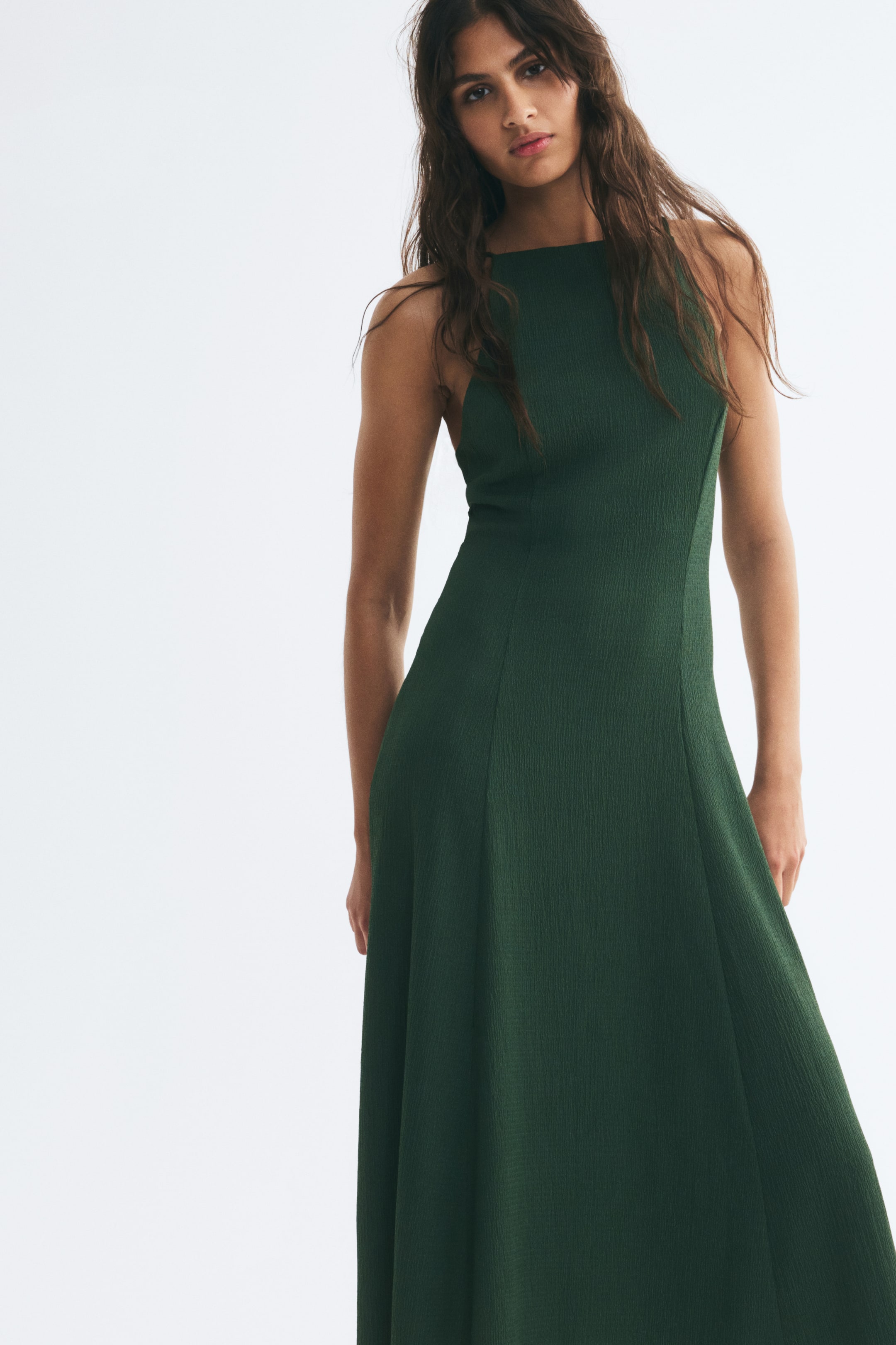 View larger image: The model is wearing a dark green crinkled jersey dress featuring a high square neckline and a fitted bodice with visible vertical seams. The dress flows into a flared skirt, highlighting the textured fabric and panel construction.