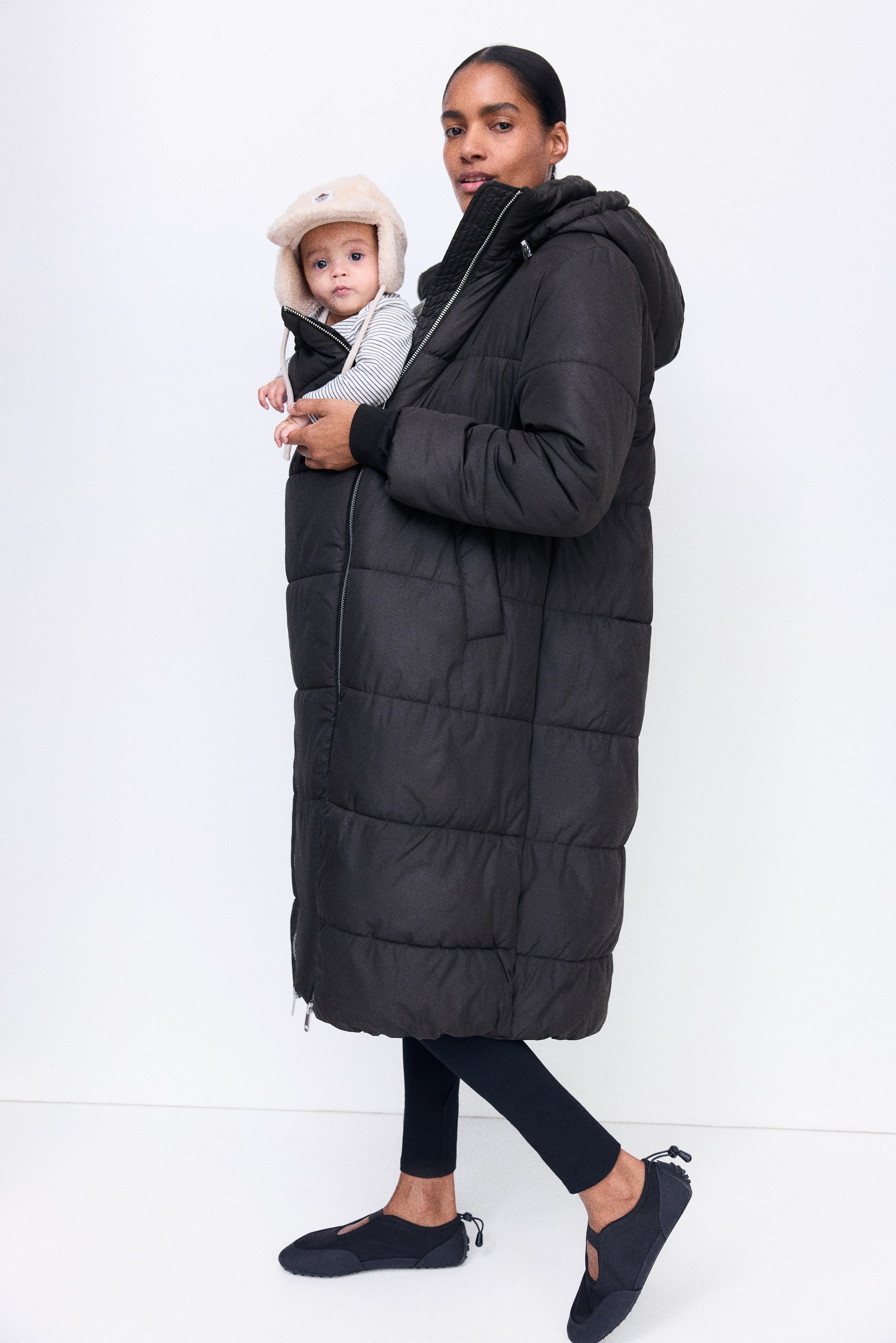 Ampliar la imagen: La chamarra MAMA Before & After Babywearing, un abrigo largo acolchado tipo puffer en color negro, es llevada por la modelo, quien carga a un bebé dentro del panel frontal de extensión con cierre. La modelo sostiene al bebé con una mano mientras usa leggings negros y zapatos negros tipo slip-on.