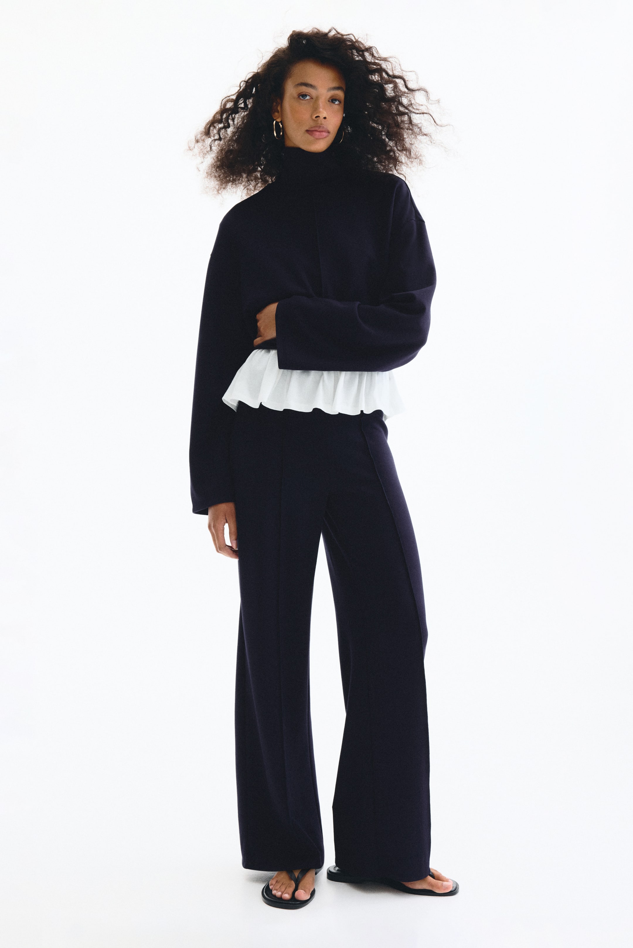 View larger image: A navy two-piece outfit is showcased from the front. The top is a cropped turtleneck sweater with wide sleeves, and a white ruffled layer is visibly peeking out from its hem. The matching pants are wide-leg and full-length.