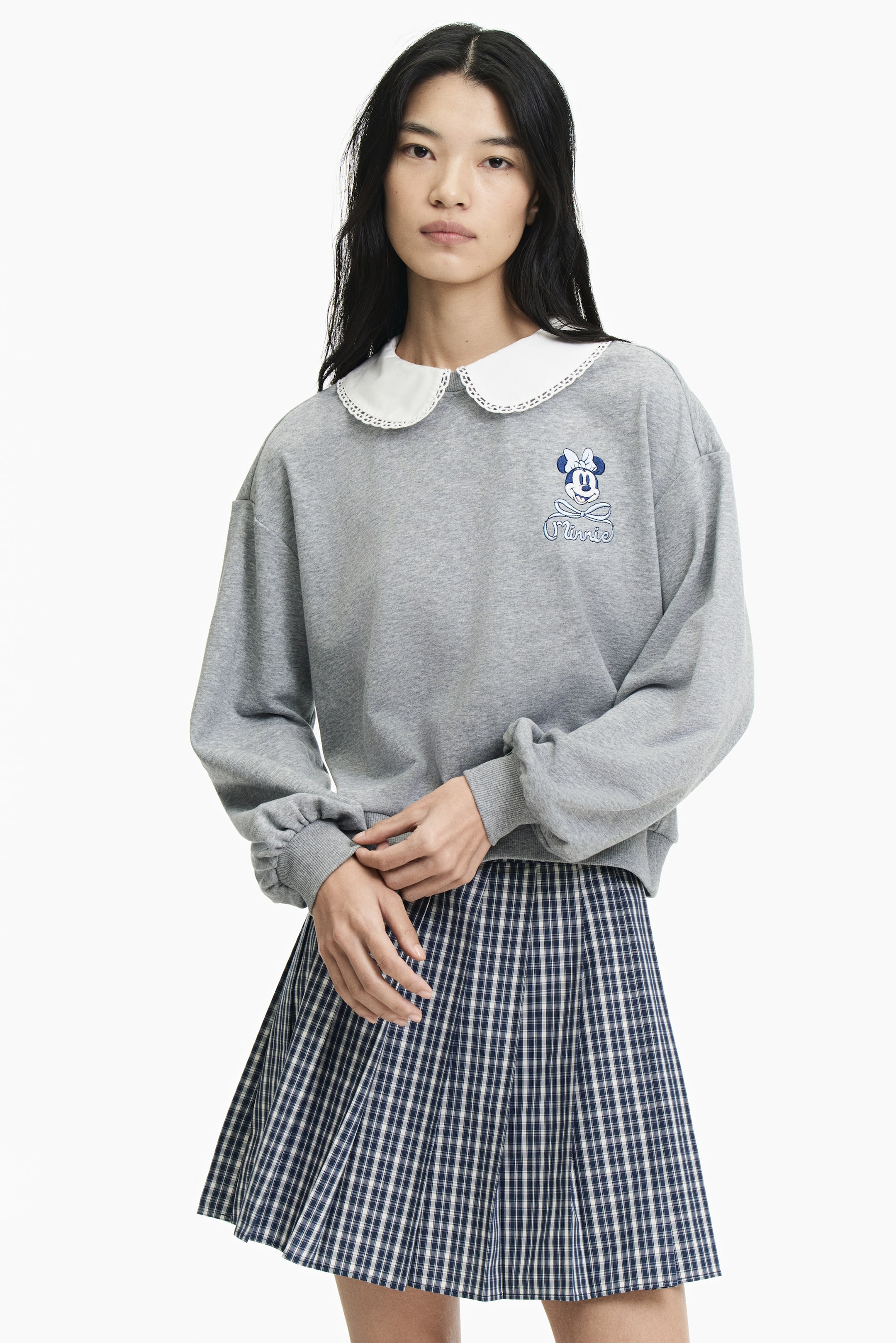 Xem ảnh lớn hơn: Người mẫu đang mặc một chiếc áo top màu xám bằng chất liệu sweatshirt mỏng nhẹ, nổi bật với họa tiết thêu Minnie Mouse ở ngực và phần cổ sen trắng lớn viền ren. Chiếc áo có thiết kế vai rơi, tay dài phồng với bo tay dệt gân và gấu áo dệt gân, trong khi người mẫu đang đặt tay lên một chiếc chân váy xếp ly kẻ sọc.