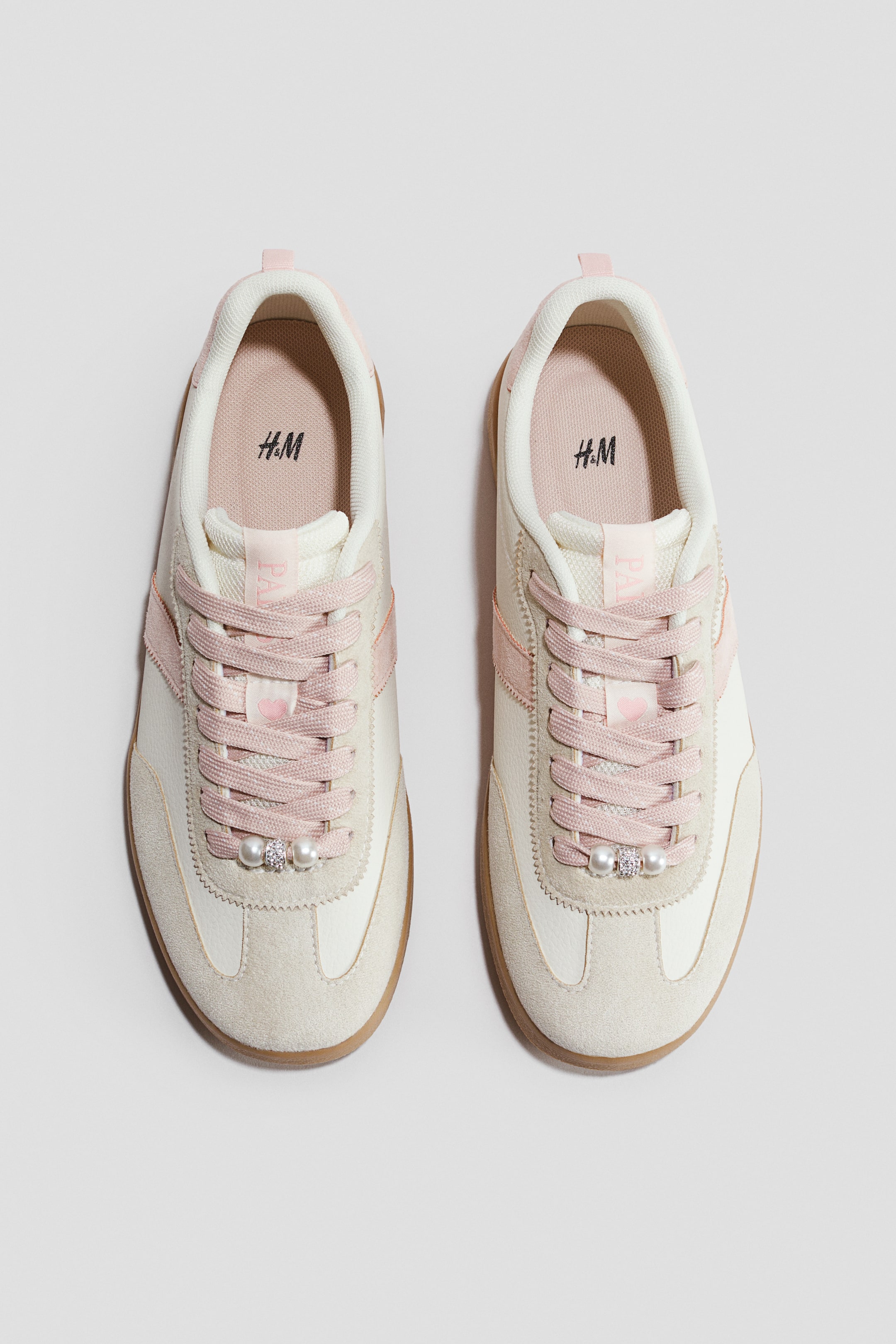 Se større billede: Et par sneakers i creme og off-white med lyserøde accenter på side-striberne og kraven. Lyserøde snørebånd krydser over tungen, og hvert snørebånd prydes af en perle og en rhinstenperle.
