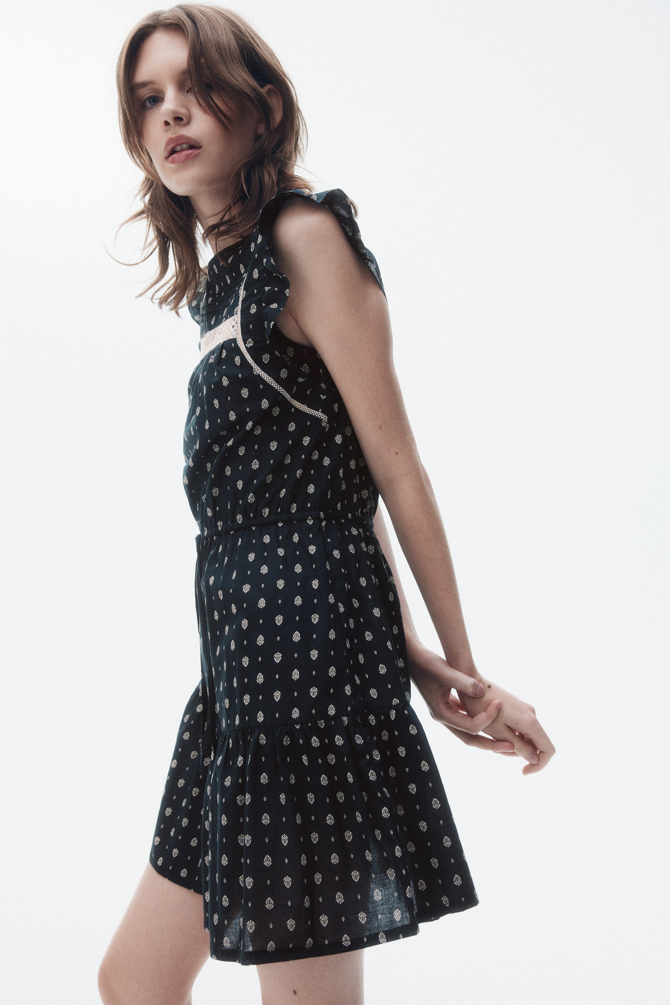 Xem ảnh lớn hơn: Một chiếc váy màu tối, nhìn từ góc nghiêng, có họa tiết lặp lại với họa tiết lá màu sáng. Nó có tay áo nhún bèo, chi tiết ren tinh tế ở phần ngực phía trước, eo co giãn, và chân váy mini phân tầng.