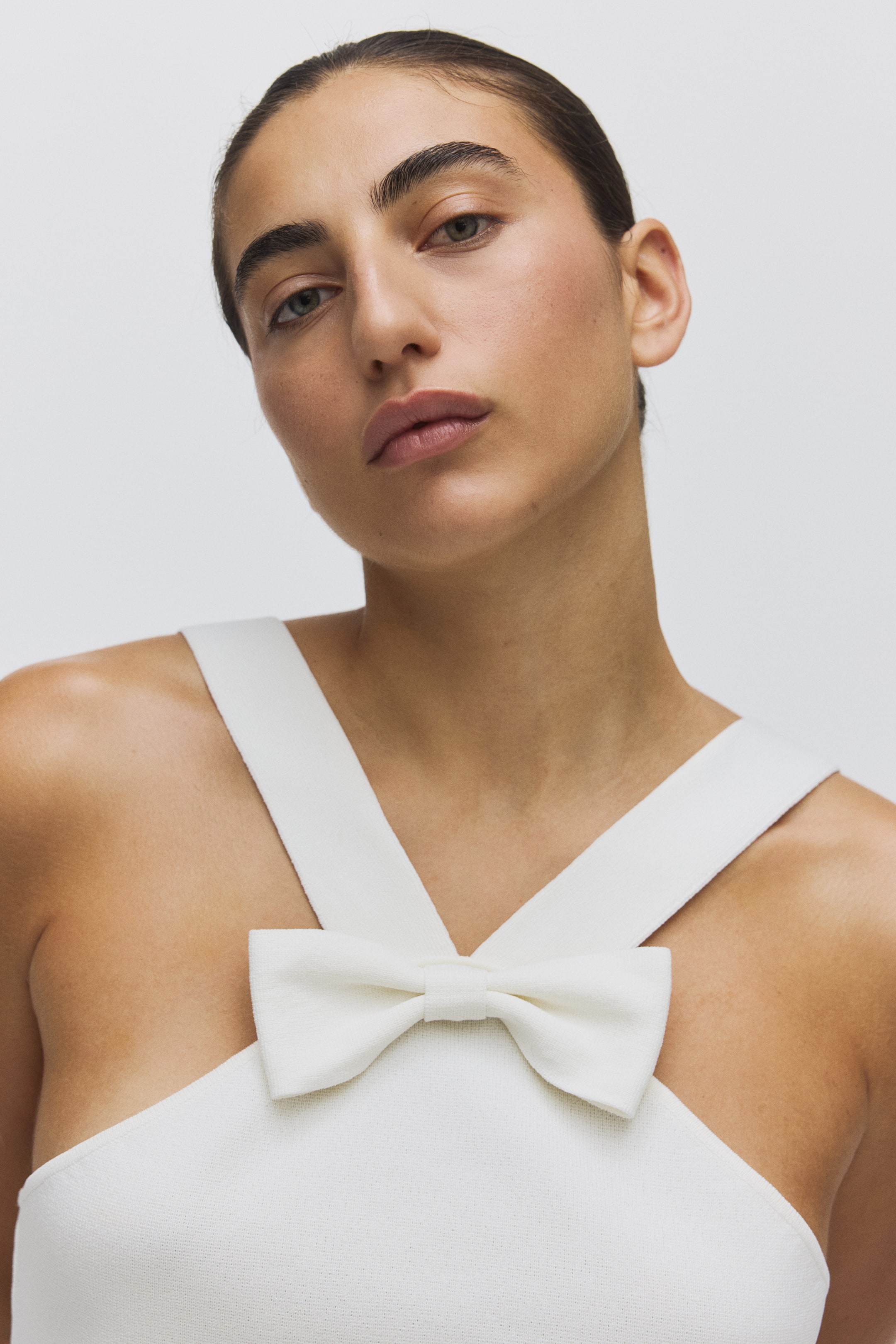 View larger image: A white garment with a subtle texture features wide straps crossing over the chest, where a textured bow is centrally positioned. The product is viewed from a front-facing, slightly angled perspective, highlighting its design.
