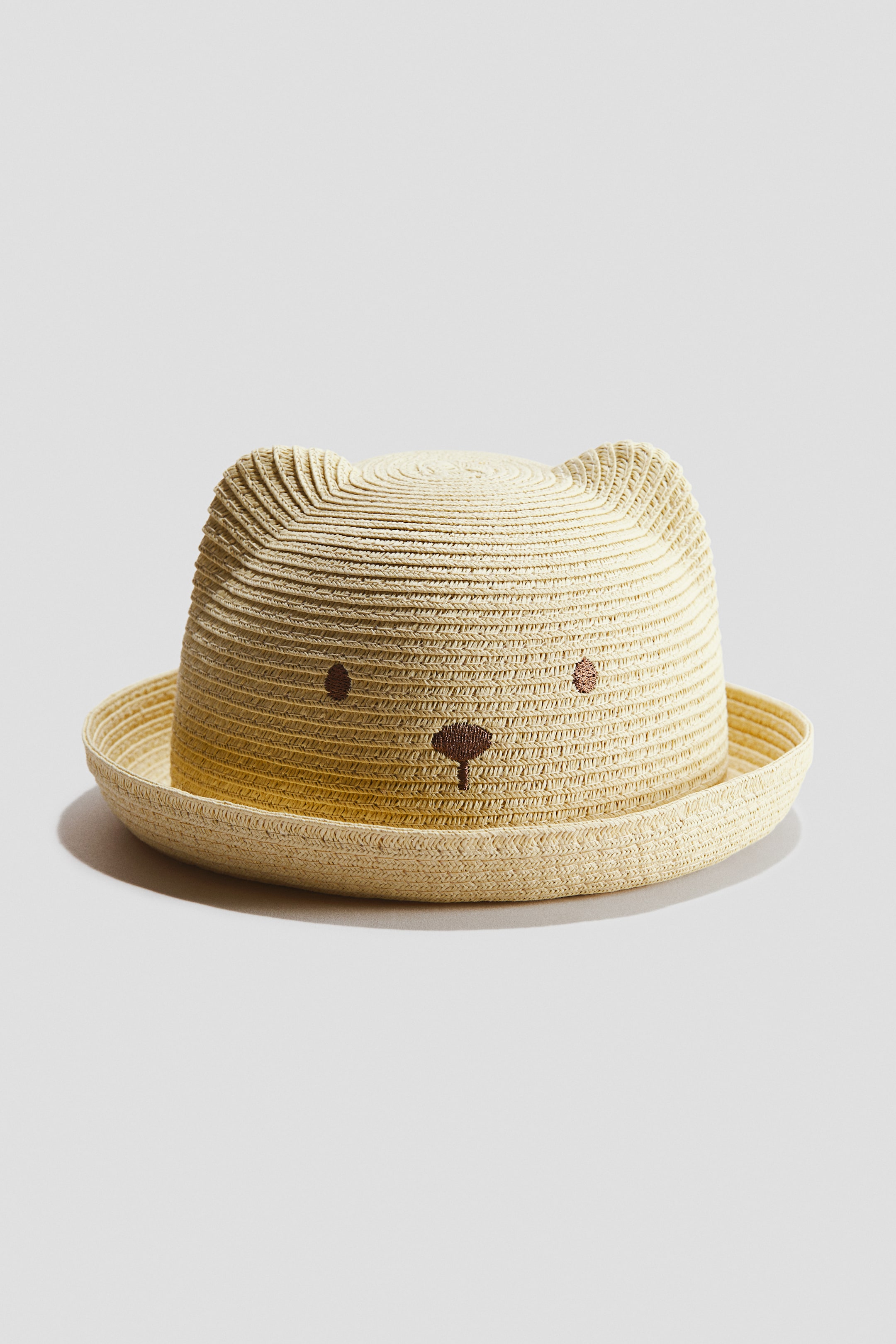 View larger image: A light beige woven straw hat showcases a rounded crown adorned with two distinct ear-like shapes on top and a flat, short brim. The front of the crown features embroidered brown dots representing eyes and a small, inverted brown triangle for a nose, forming an animal face.