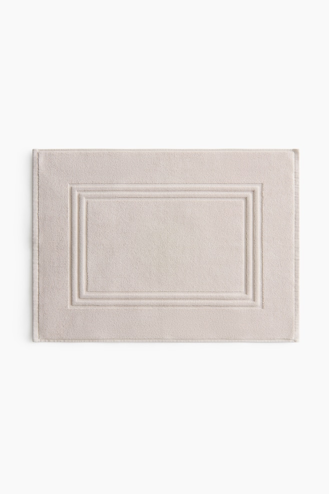 A rectangular bath mat in a light beige cotton terry is showcasing its front. It boasts a subtle sheen and a raised, rectangular frame detailing its center, with a woven trim completing its outer edges. - 1