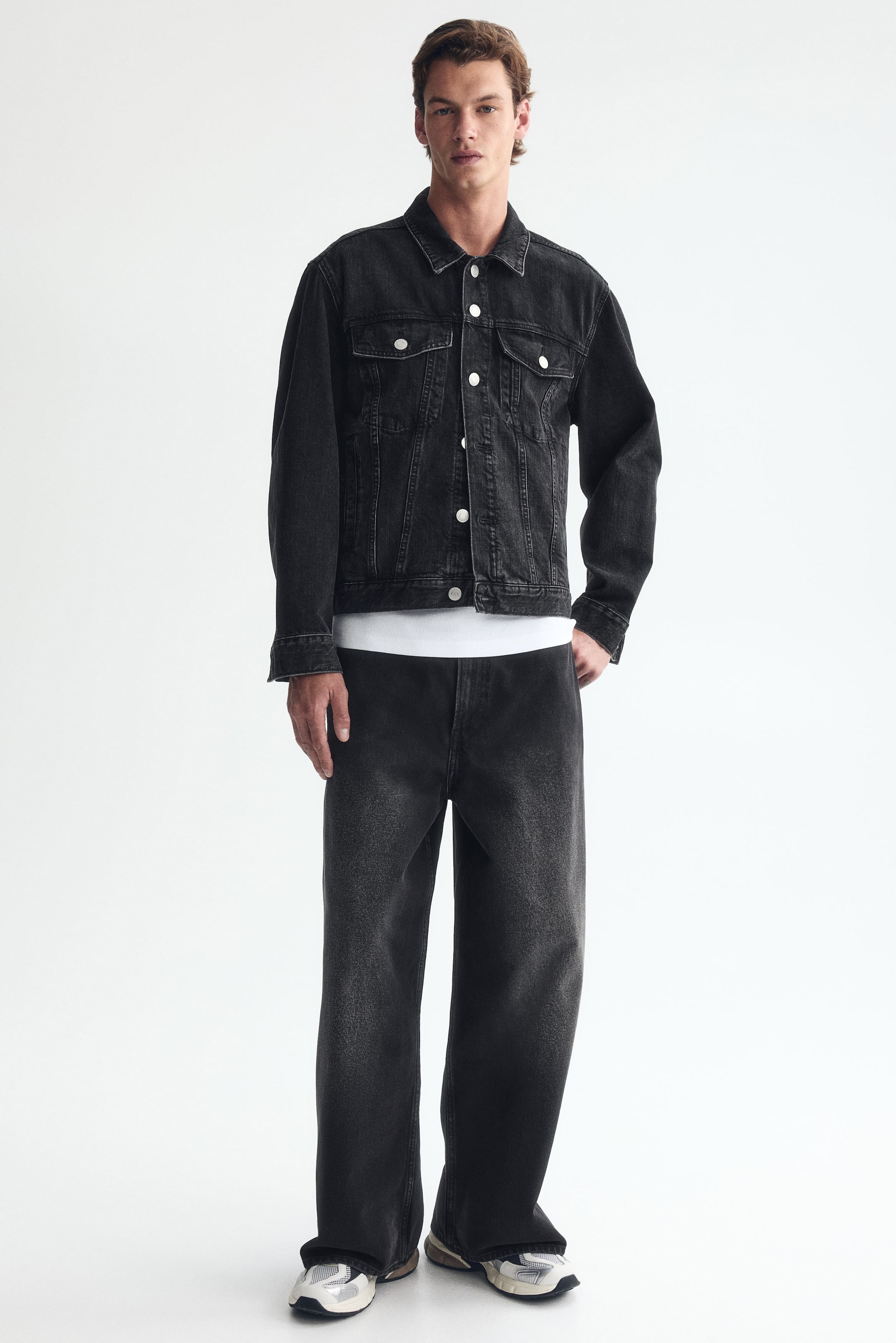 View larger image: The model is wearing 5-pocket jeans crafted from dark wash rigid cotton denim. These jeans boast a regular waist, a dropped crotch, and a rounded, baggy silhouette that provides ample room throughout the entire leg, elegantly stacking at the ankle over white sneakers.