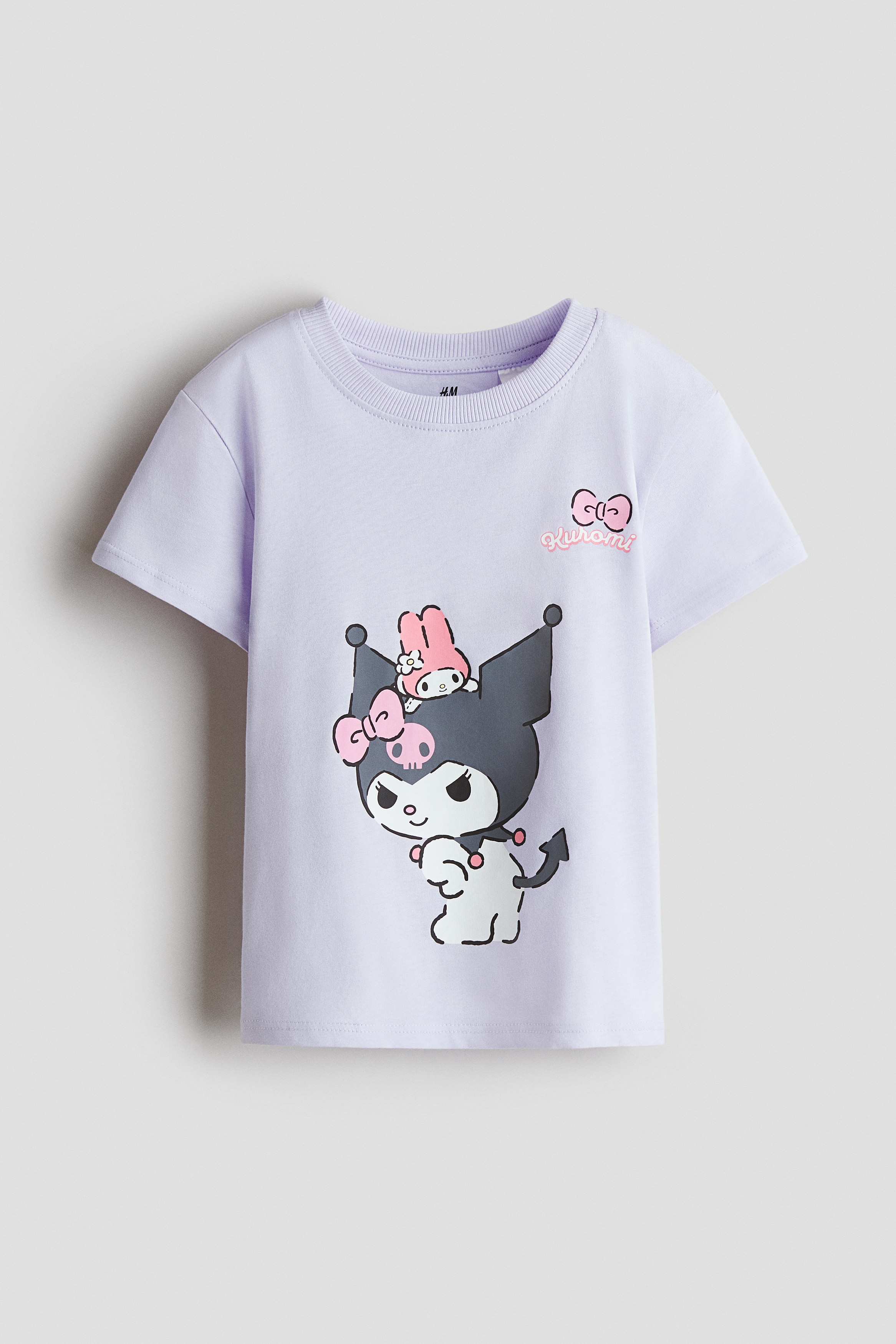 A light purple cotton T-shirt featuring a ribbed round neckline, dropped shoulders, and a graphic character print at the front is worn by the model. The T-shirt is styled with light-wash denim jeans.