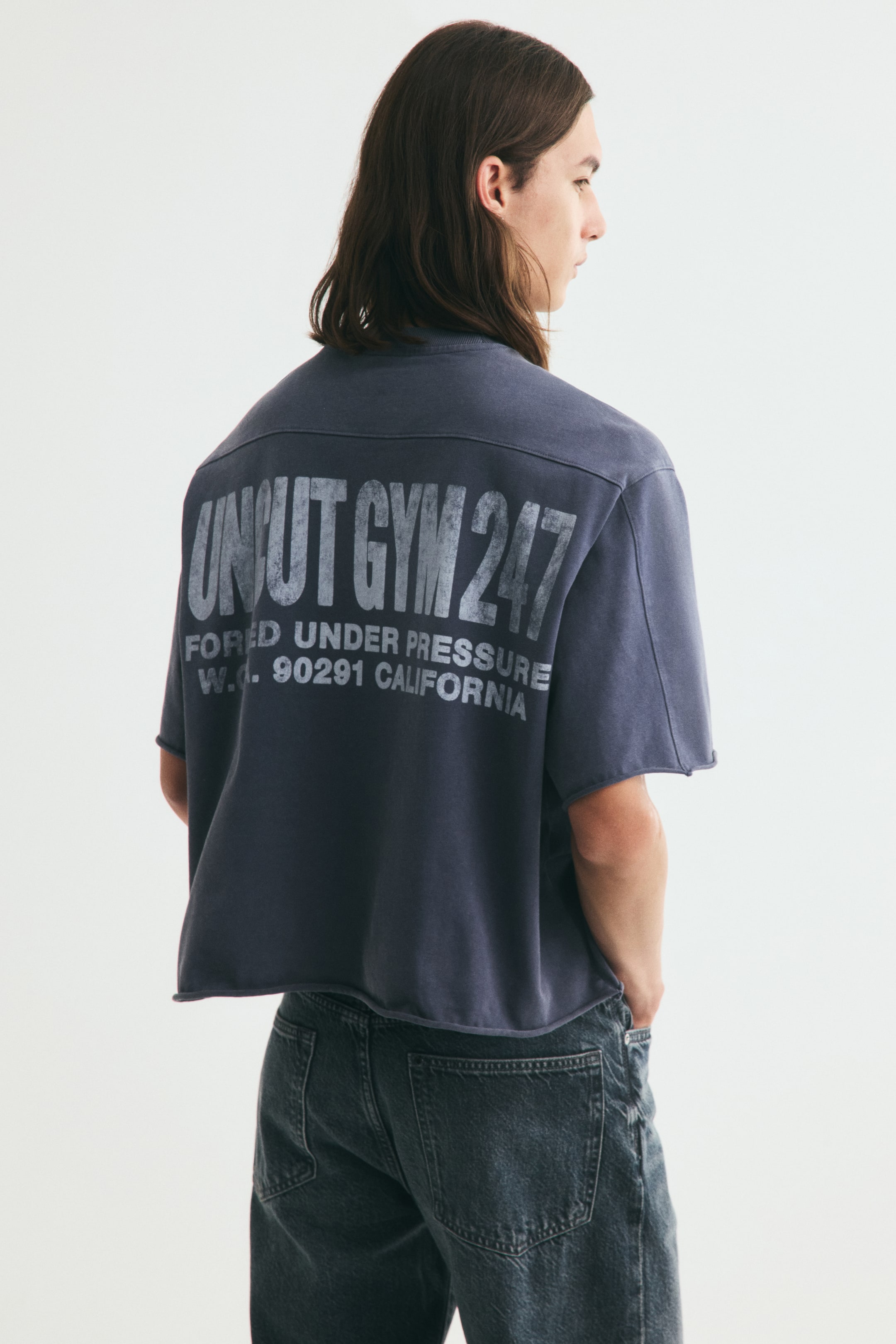 View larger image: A short, dark gray, boxy-style T-shirt in heavyweight cotton jersey is worn by the model, featuring a round, rib-trimmed neckline and dropped shoulders. The back displays a distressed white print reading "UNCUT GYM 247" and "FORCED UNDER PRESSURE W.C. 90291 CALIFORNIA", while rolled edges finish the cuffs and hem, creating an oversized, baggy silhouette.
