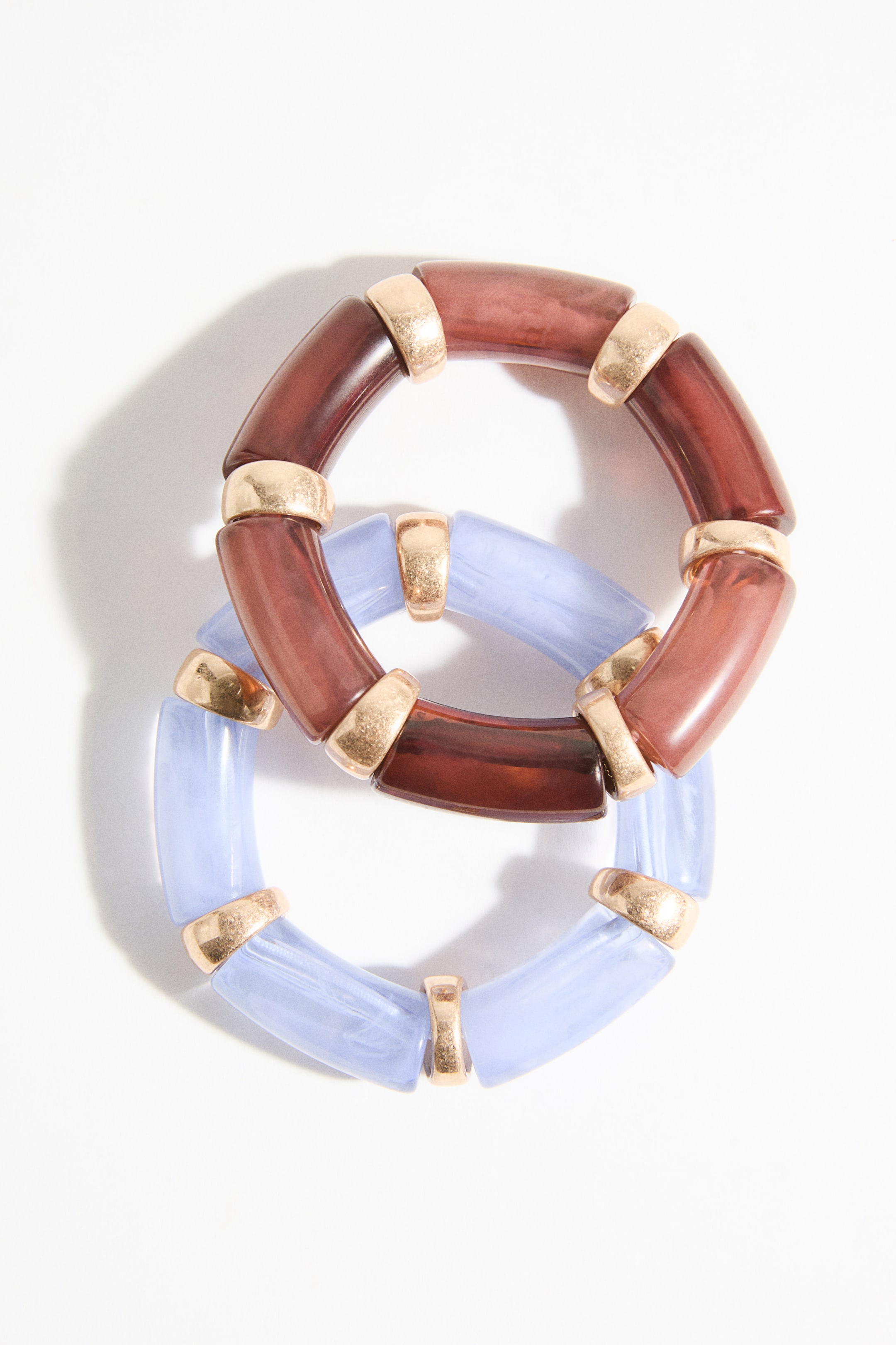 Zobraziť väčší obrázok: Dva náramky sú prepojené, tvoria tvar osmičky. Jeden náramok zobrazuje priesvitné červeno-hnedé zakrivené segmenty, zatiaľ čo druhý zobrazuje priesvitné svetlo modré zakrivené segmenty, všetky spojené textúrovanými, zlatohnedými kovovými krúžkami.