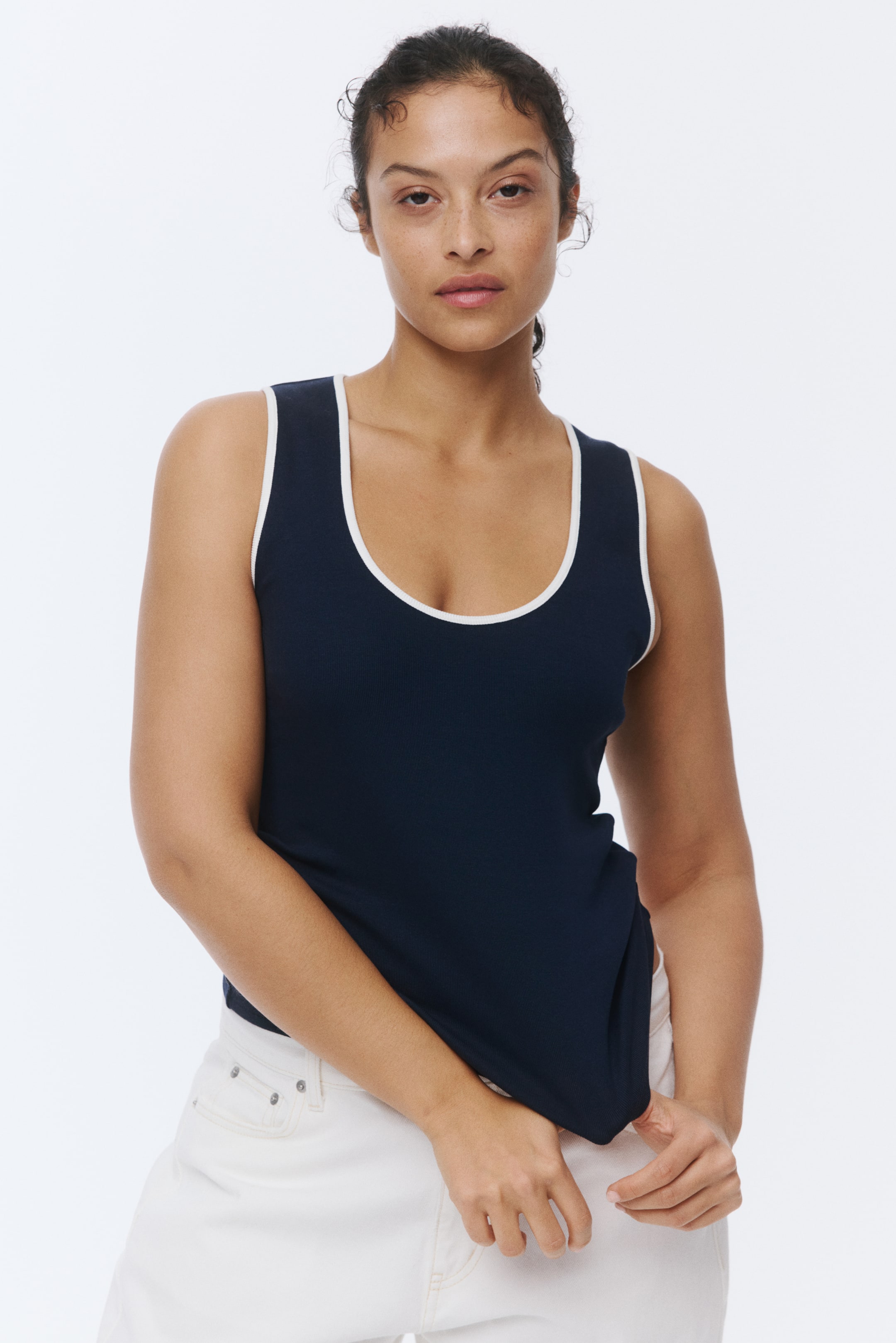 View larger image: The model is donning a navy fitted vest top crafted from ribbed cotton jersey. It boasts a scoop neckline, a straight-cut hem, and contrasting white trims around the neckline and armholes. The model is seen holding the lower edge of the top.