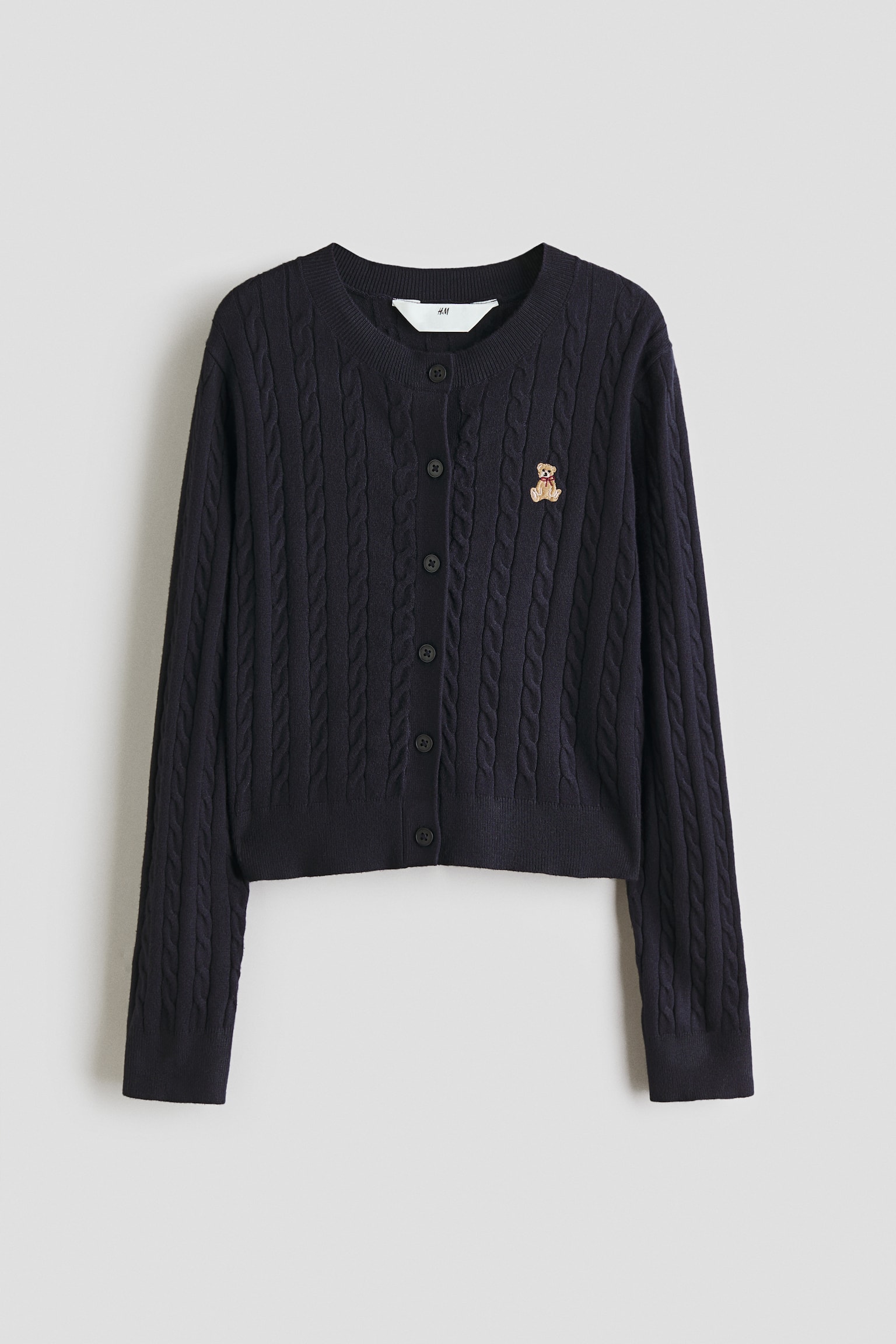 A dark blue cable knit cardigan is lying flat, front-facing, featuring a round, rib-trimmed neckline and six buttons down the front. A small embroidered teddy bear detail is appearing on the left chest, while long sleeves with ribbed cuffs and a ribbed hem are completing the design. - 1