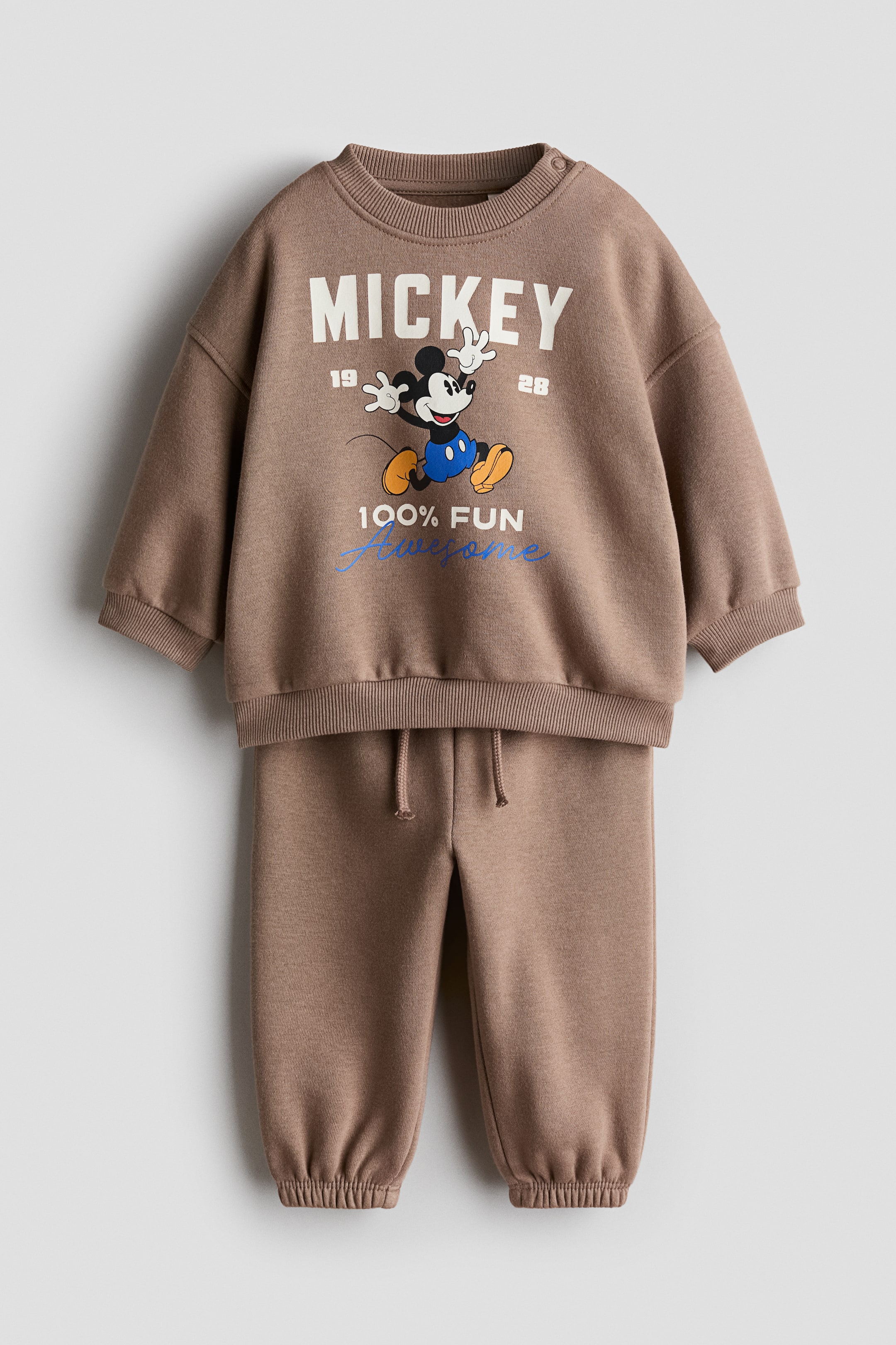 View larger image: A light brown 2-piece printed sweatshirt set is showcasing its front. The crewneck sweatshirt features shoulder snap buttons, ribbed cuffs and hem, and a graphic print of Mickey Mouse with the words 'MICKEY', '19', '28', and '100% FUN Awesome'. The matching sweatpants come with a drawstring waist and ribbed ankle cuffs.