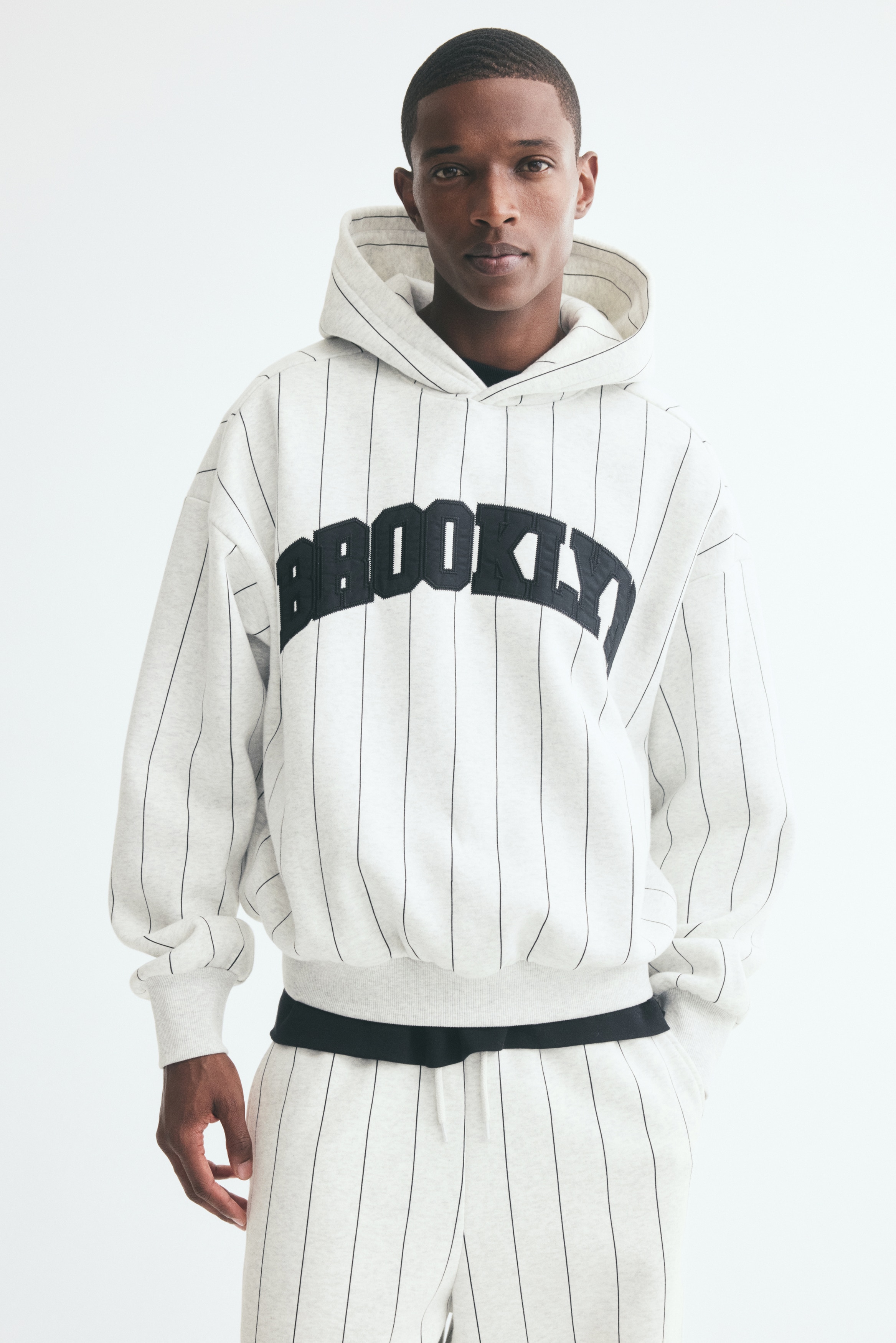 Má»t chiáº¿c hoodie mÃ u xÃ¡m nháº¡t káº» sá»c máº£nh Äang ÄÆ°á»£c chá»¥p trá»±c diá»n, ná»i báº­t vá»i dÃ²ng chá»¯ "BROOKLYN" dáº¡ng chá»¯ varsity mÃ u Äen uá»n cong ngang ngá»±c. Sáº£n pháº©m cÃ³ thiáº¿t káº¿ vai trá», tay dÃ i vá»i bo tay gÃ¢n, gáº¥u Ã¡o gÃ¢n vÃ  mÅ© chá»ng lá»p chÃ©o.