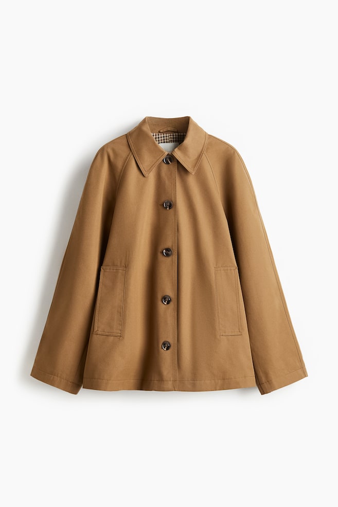 Eine braune Jacke wird frontseitig angezeigt, mit einem spitzen Kragen und einem sichtbaren karierten Futter. Sie ist mit Raglanärmeln, einem Knopfverschluss mit fünf braunen Knöpfen und zwei großen aufgesetzten Vordertaschen gestaltet. - 1