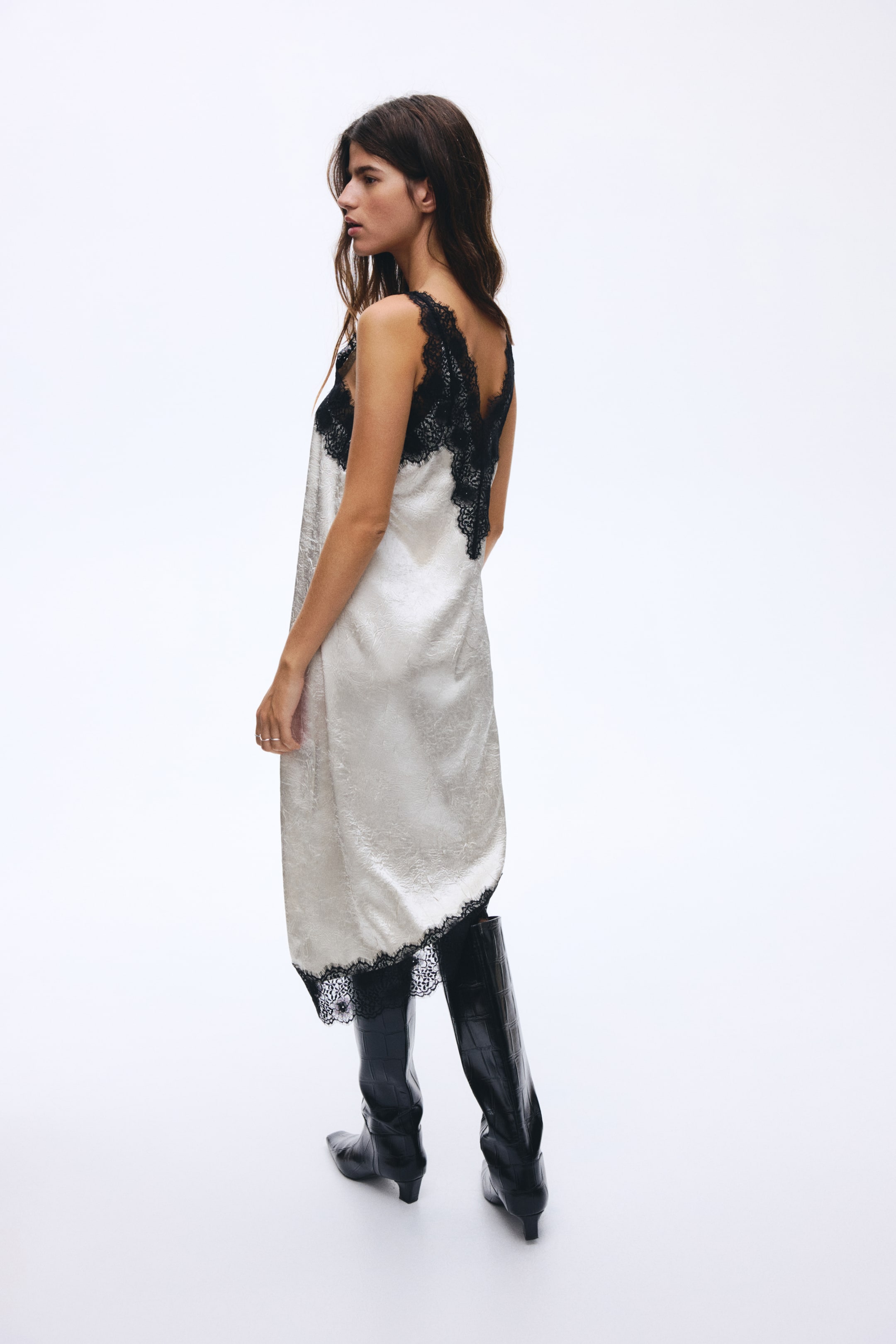 Zobraziť väčší obrázok: Lesklé, bielo-krémové šaty bez rukávov s jemným vzorom pripomínajúcim damask sú naklonené na trištvrte doľava a trochu dozadu. Predstavujú čierne čipkové lemovanie pozdĺž výstrihu v tvare V, ktoré sa rozširuje cez plecia a tvorí hlboký V na chrbte, a ohraničujú stredne dlhé lemovanie.