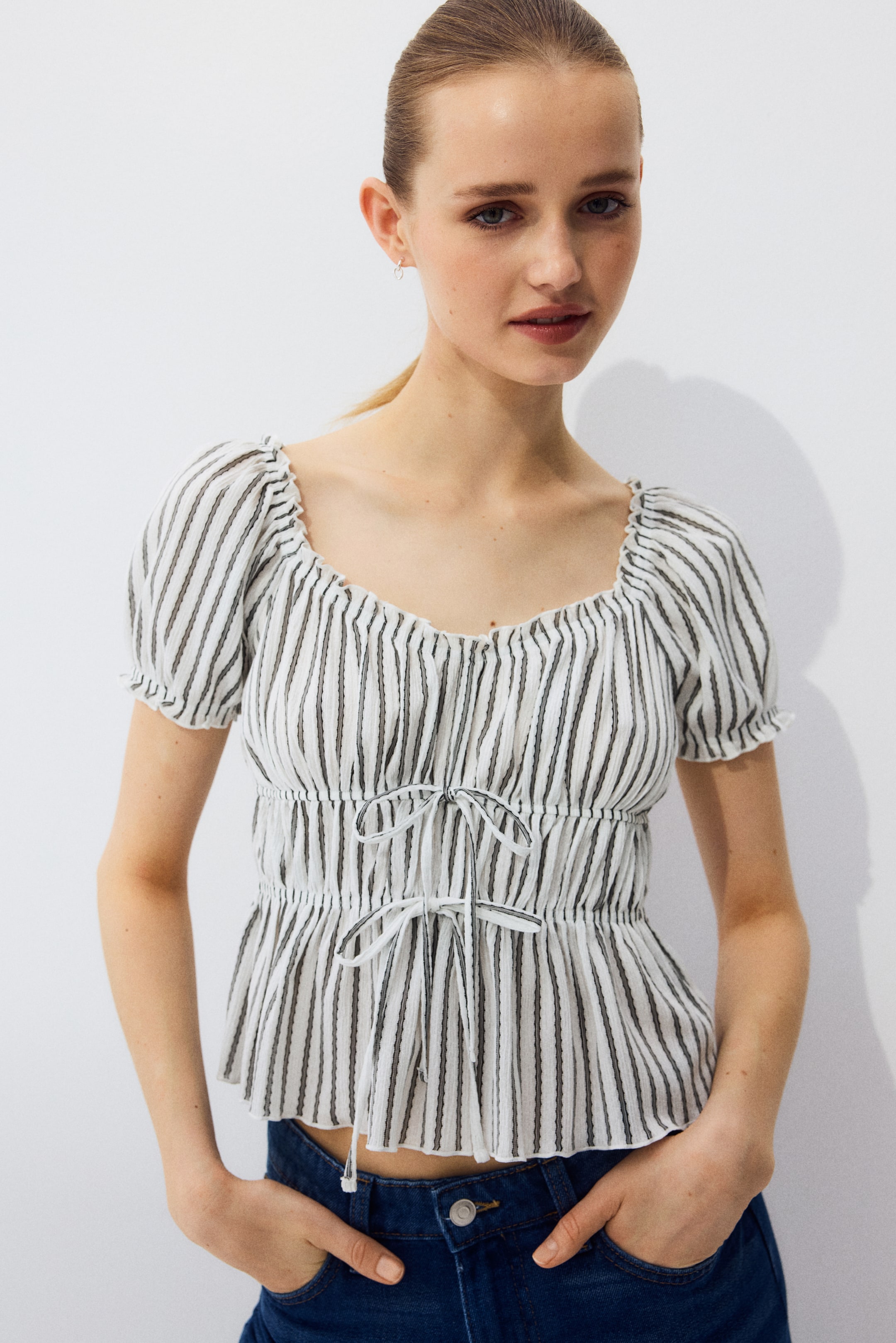 View larger image: A white top adorned with slender black vertical stripes showcases a gathered sweetheart neckline accompanied by short puffed sleeves. The waist is cinched with smocked detailing, while two white ties featuring black stripes are elegantly knotted at the front, forming a peplum hem.
