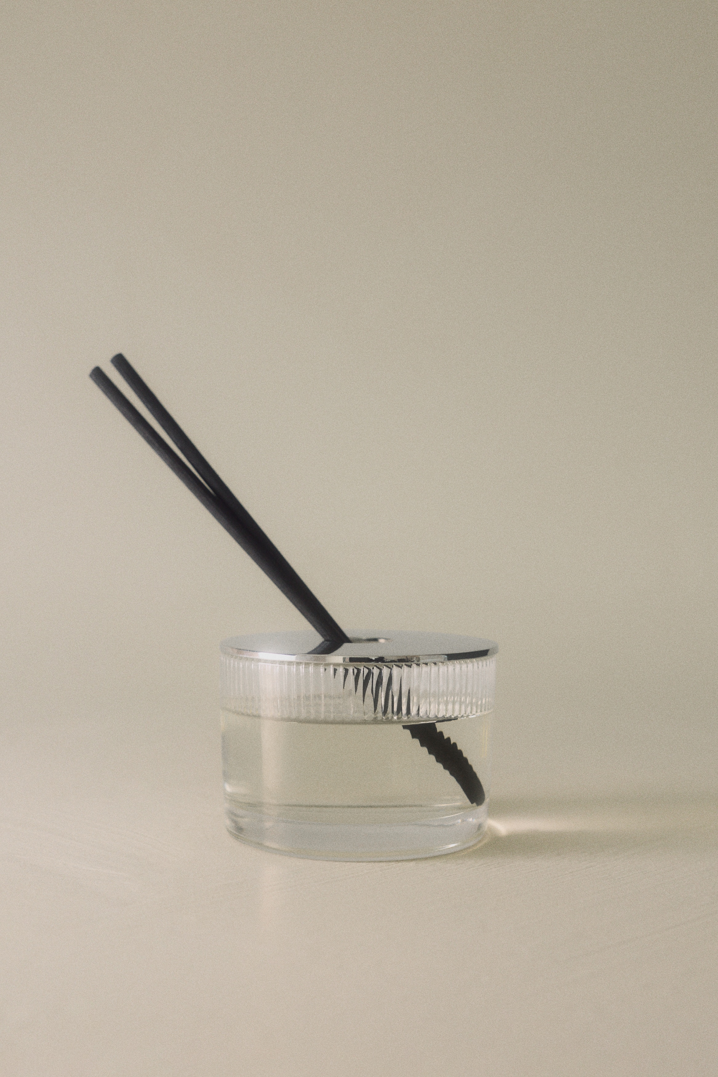 Een geurdiffuserset wordt van voren getoond en bestaat uit een witte gestructureerde doos met in reliÃ«f aangebrachte âDARK PATCHOULIâ-tekst aan de linkerkant, een doorzichtige plastic navulfles met lichtgele vloeistof in het midden, en een geribbelde doorzichtige glazen houder met een gepolijste zilverkleurige dop met vijf zwarte diffuserstokjes aan de rechterkant.