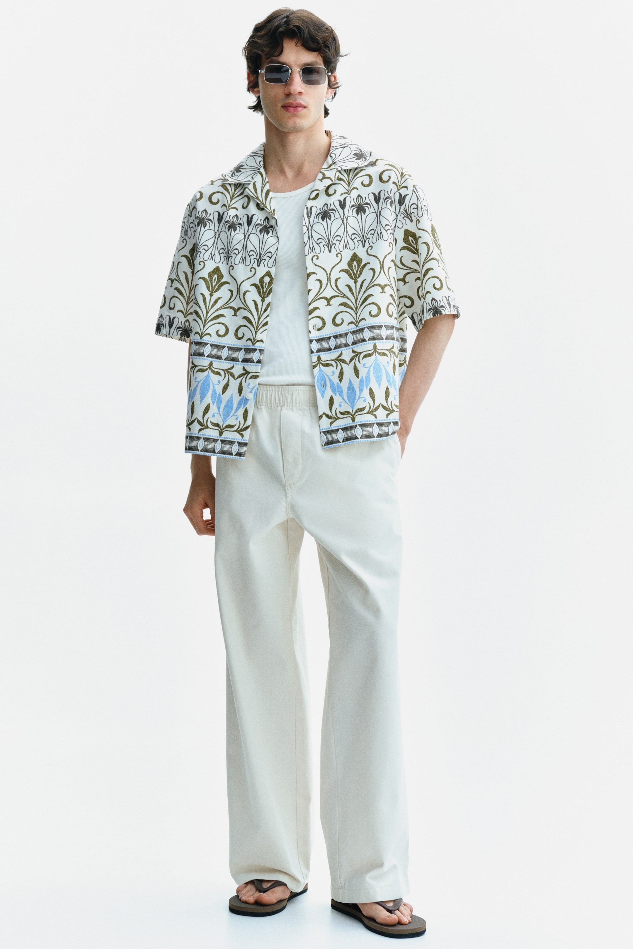 View larger image: The model is sporting a camp collar short-sleeve shirt with an intricate olive green, black, and light blue botanical pattern, complemented by horizontal bands on an off-white backdrop. The shirt is styled unbuttoned, layered over a white ribbed tank top.
