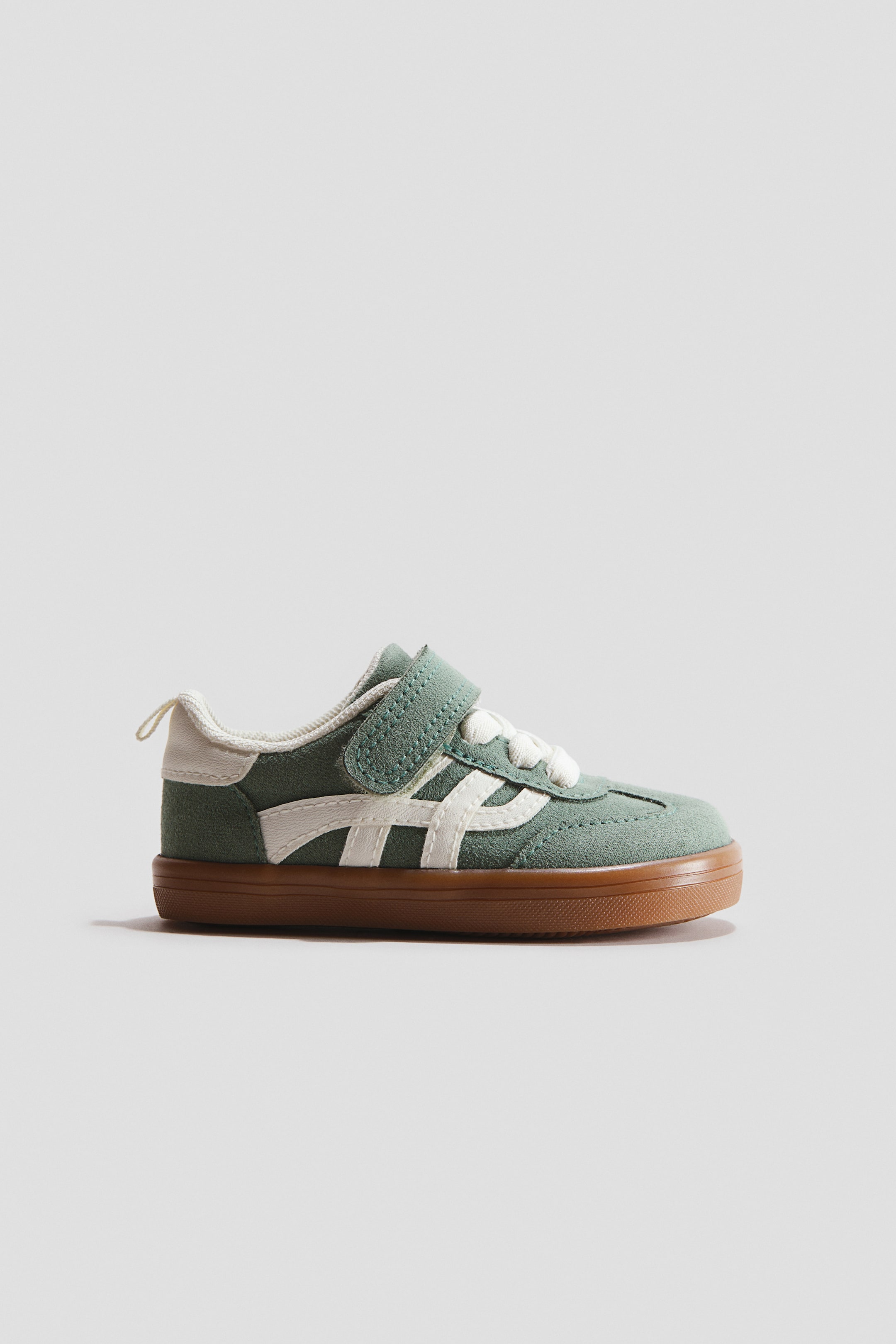 Se større billede: En børnesneaker viser sin sideprofil, med en salviegrøn ruskindsoverdel med hvide striber og en hvid polstret krave. En enkelt grøn velcrorem sikrer fronten over hvide snørebånd, mens en brun gummisål strækker sig fra tå til hæl.