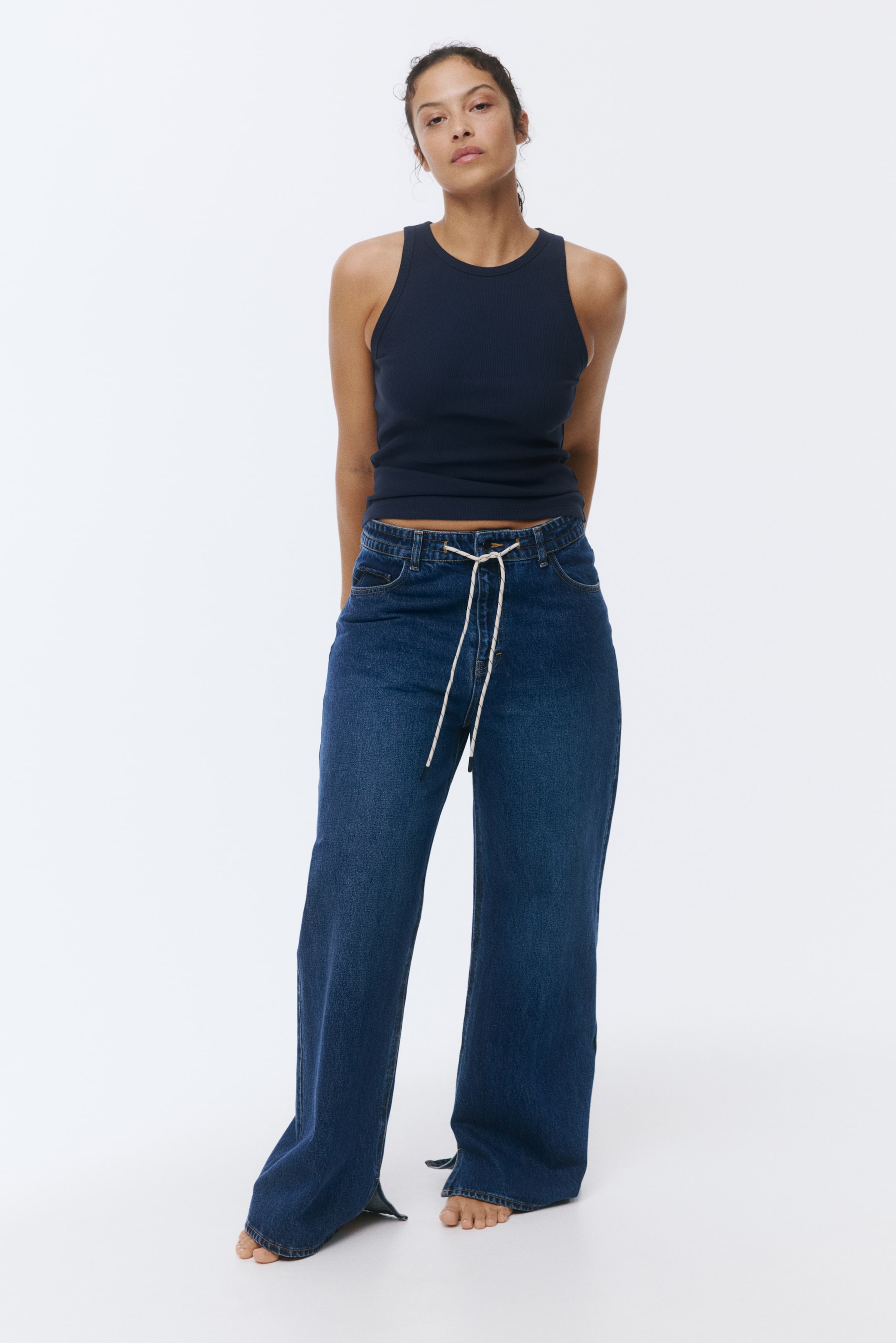 Xem ảnh lớn hơn: Áo tank top xanh đậm có gân ôm sát cơ thể và có cổ tròn. Quần jeans ống rộng màu tối có dây rút trắng ở eo và đường xẻ nhỏ ở mắt cá chân ngoài.