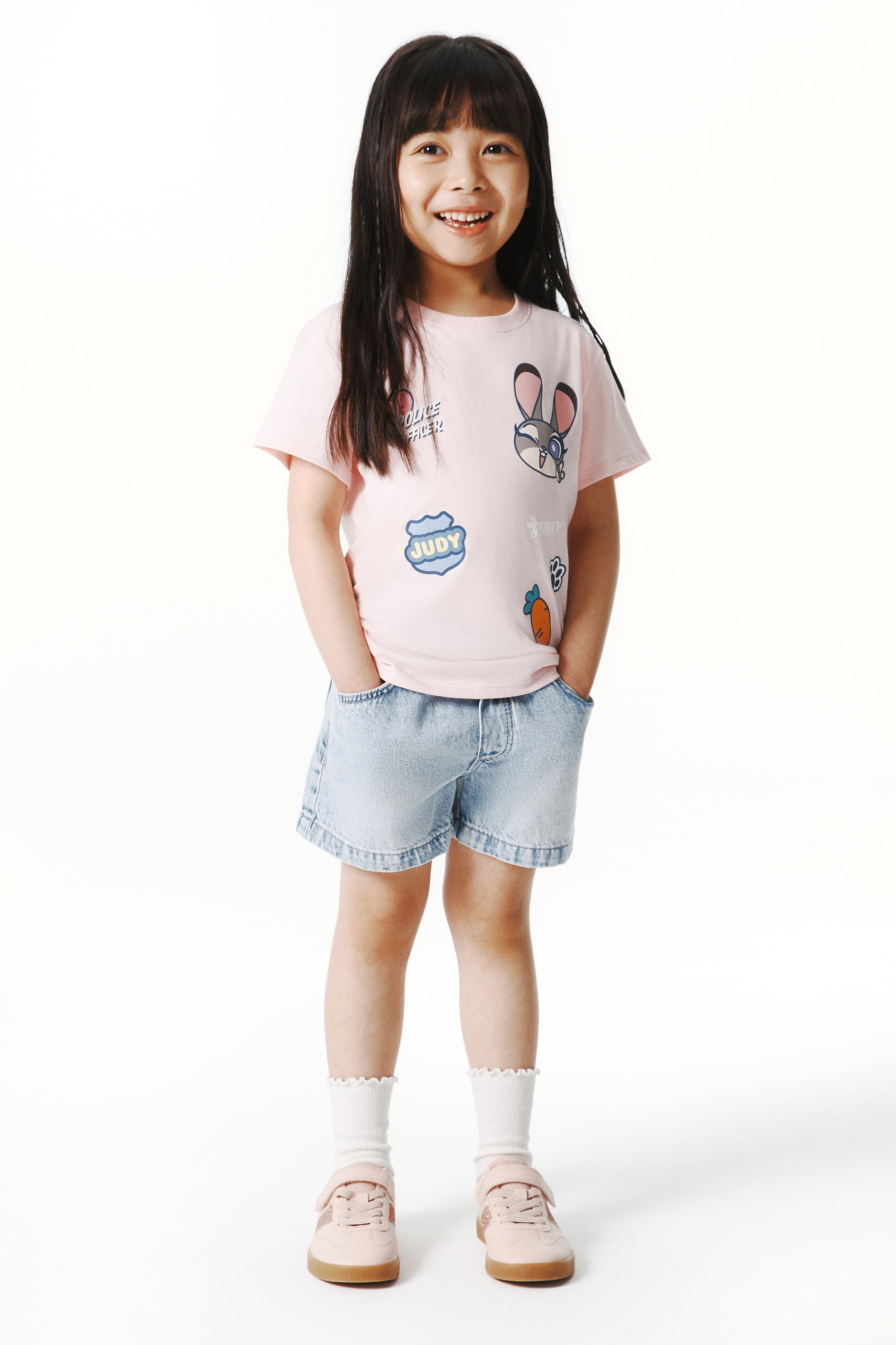 拡大画像を表示: ライトピンクの半袖Tシャツは、クルーネックで、フロントにはカラフルなパッチが複数施されています。これには、カートゥーンのウサギの顔、「POLICE OFFICER」のテキスト、「JUDY」のテキスト、そして小さなニンジンのグラフィックが含まれています。
